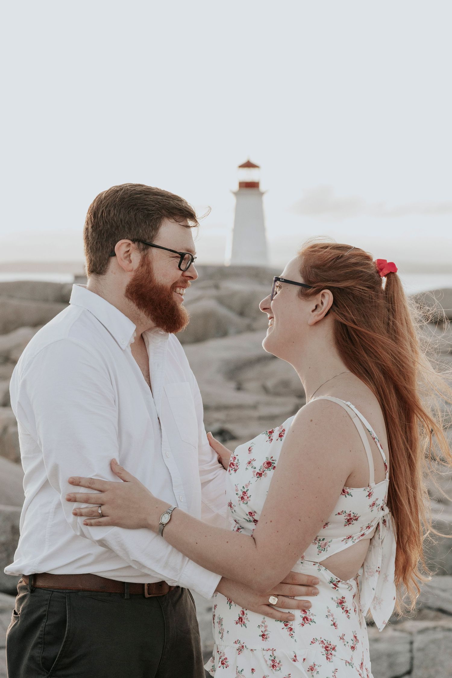 Happy couple holding each other and laughing near Peggy's Cove lighthouse on rocky coastline at sunset.