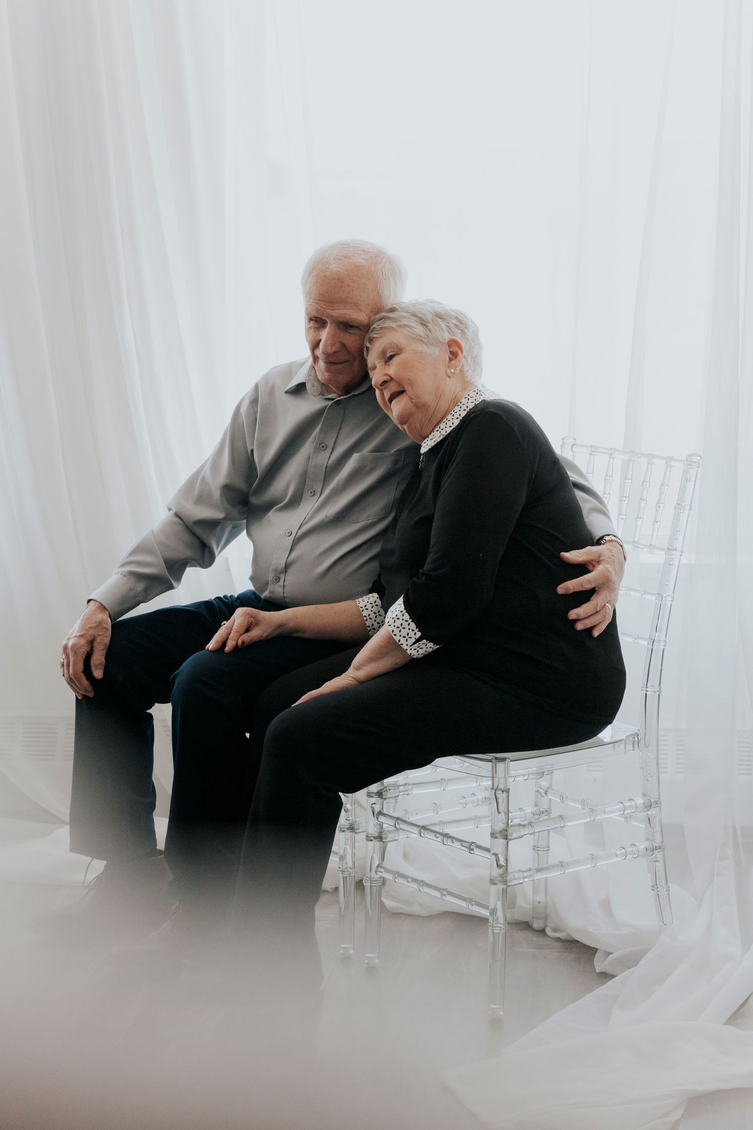 Elderly couple sitting on clear chair, sharing tender embrace against white curtain backdrop.