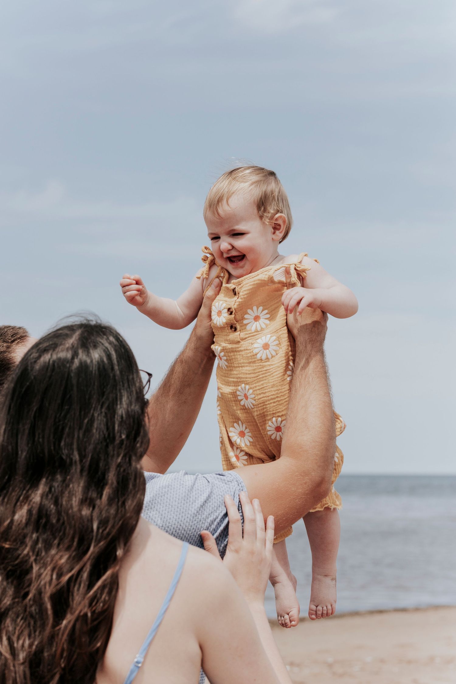 Father lifting laughing baby girl in yellow floral romper toward mother on a sunny beach.