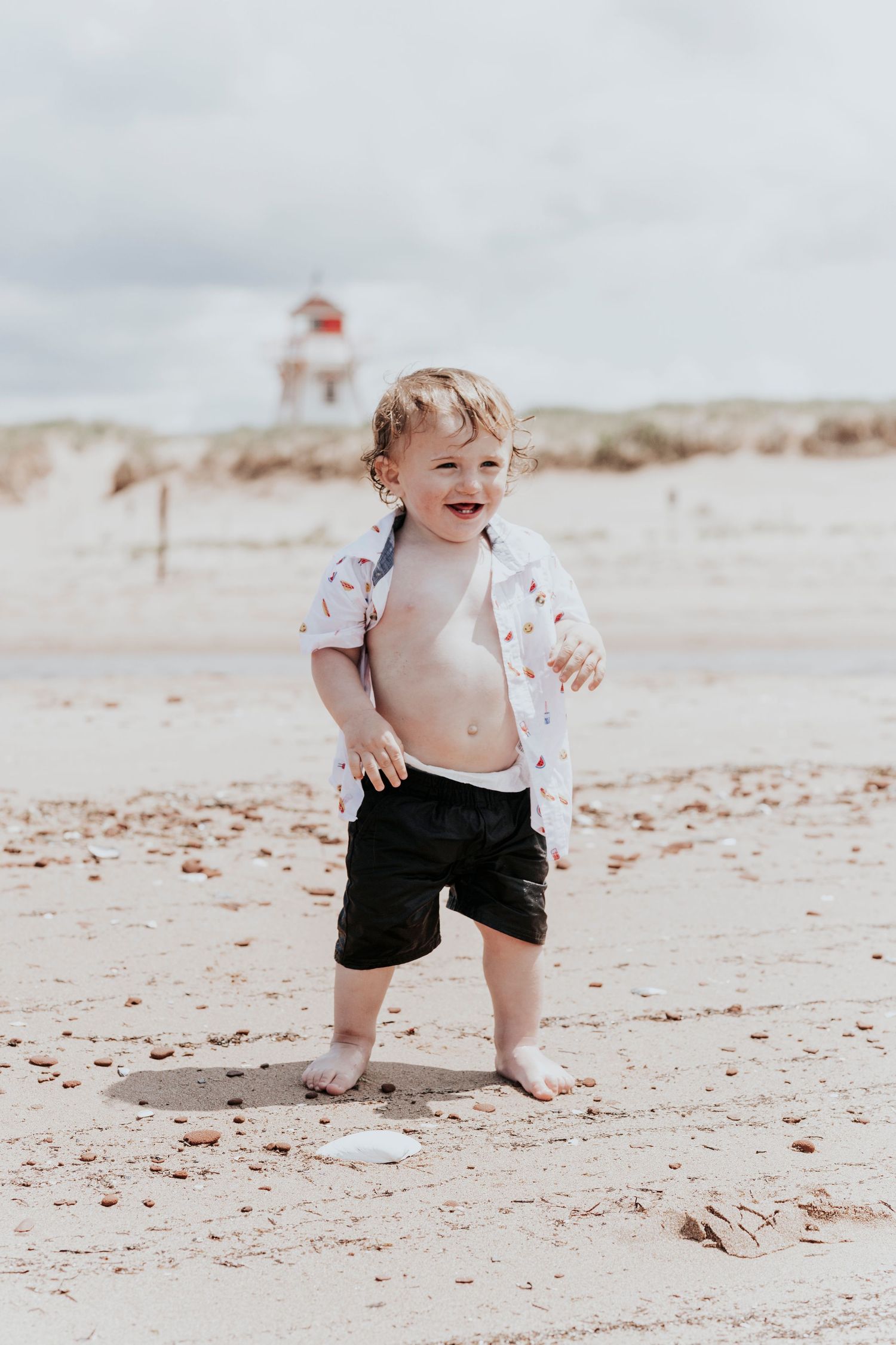 Laughing toddler boy in open shirt and black shorts playing barefoot on sandy beach with lighthouse in background.