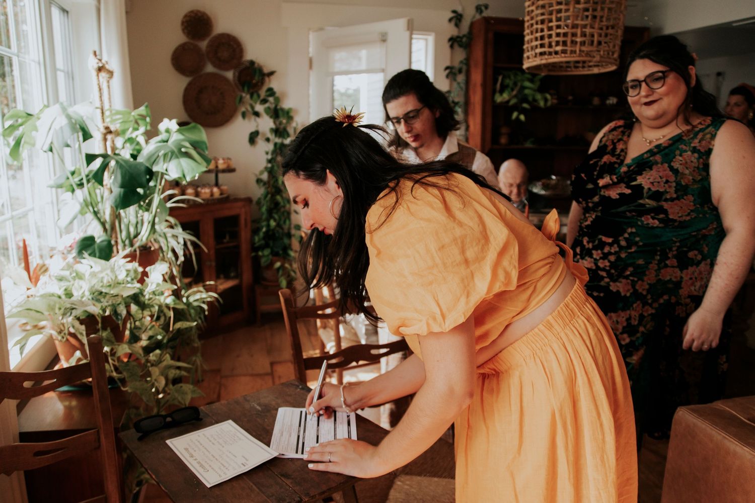 People gather in a cozy home setting around a table with houseplants and windows in warm lighting.