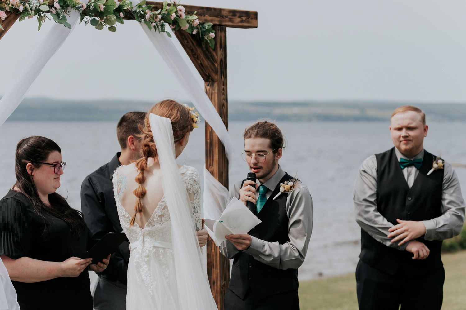 An outdoor wedding ceremony takes place under a wooden arch beside a serene waterfront on an overcast day.
