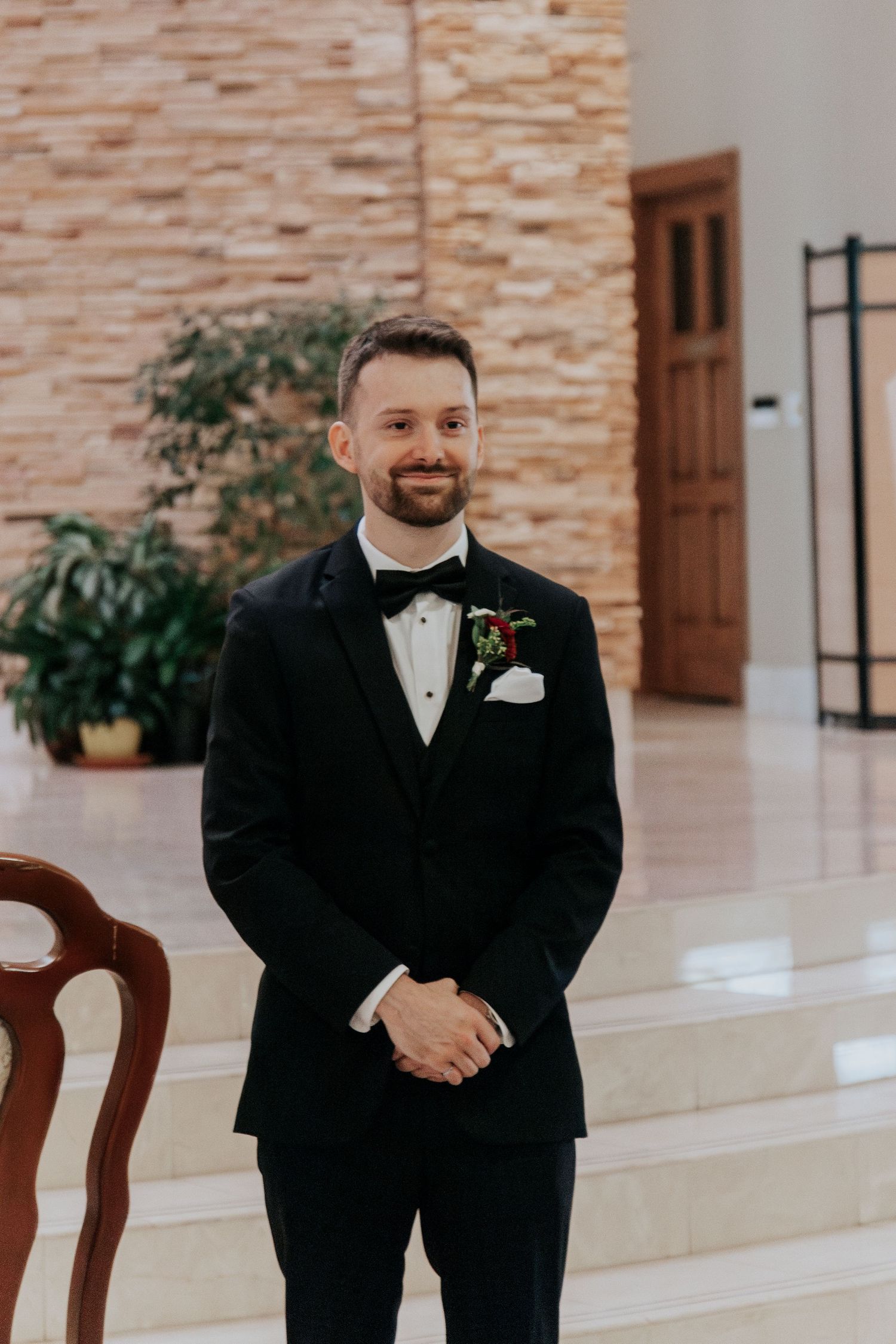 A groom in classic black tuxedo and bow tie stands in an elegant church interior.