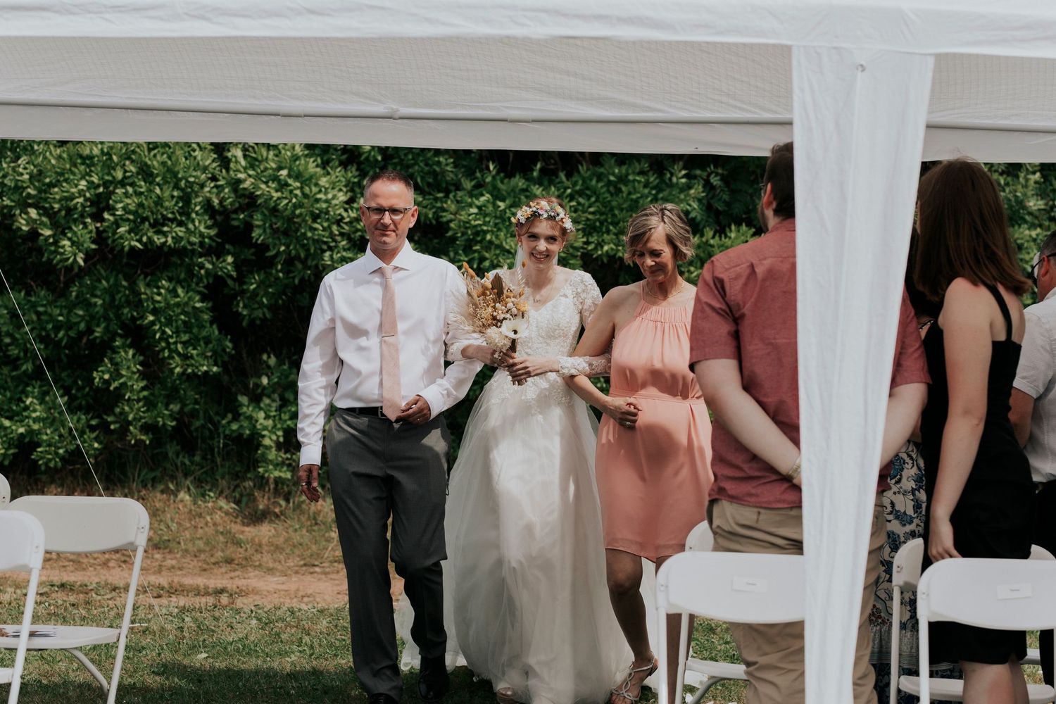 The bride and her parents make their way down the aisle on a sunny summer wedding day
