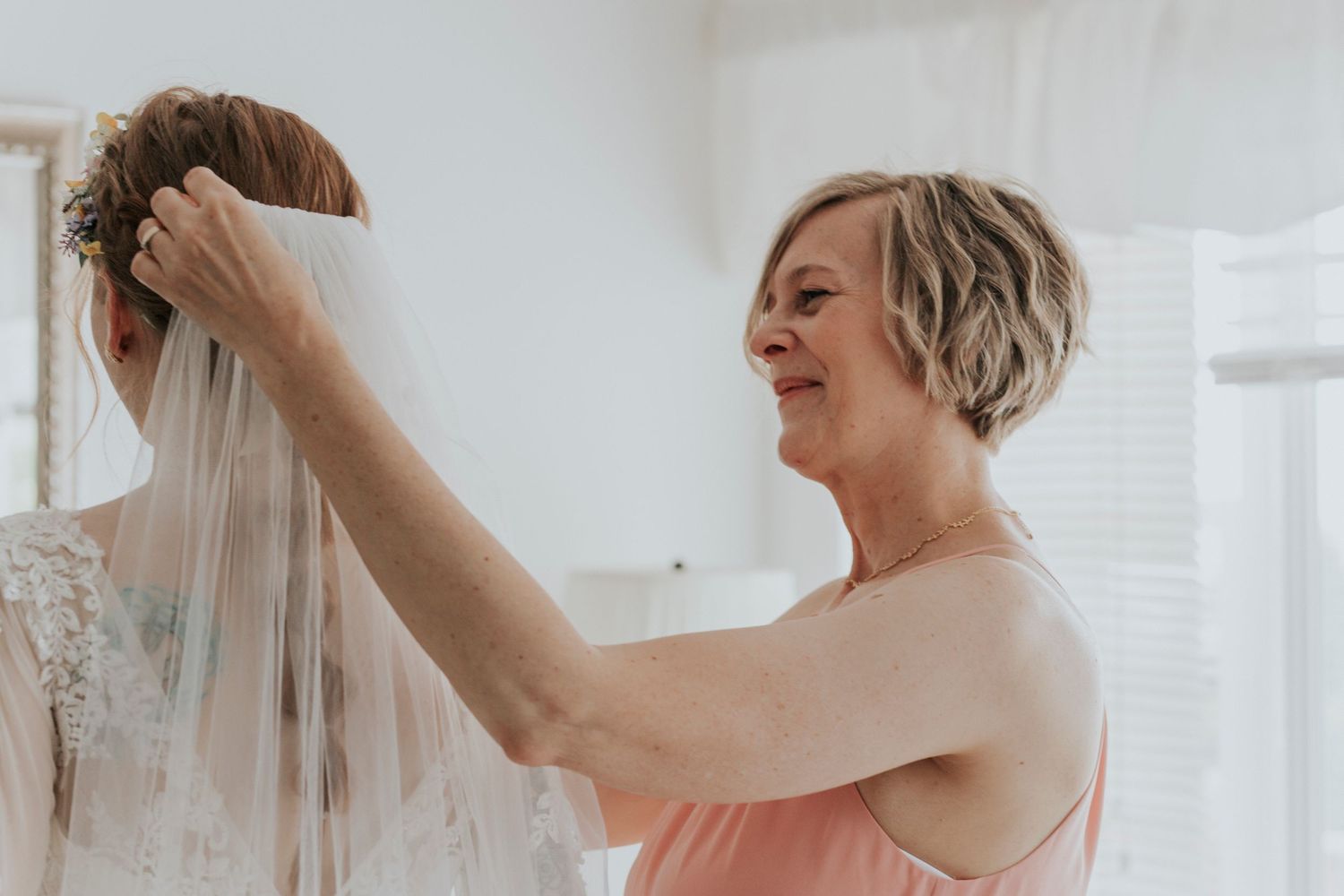 Brides mother adjusts a delicate bridal veil during wedding preparations in a bright white room.