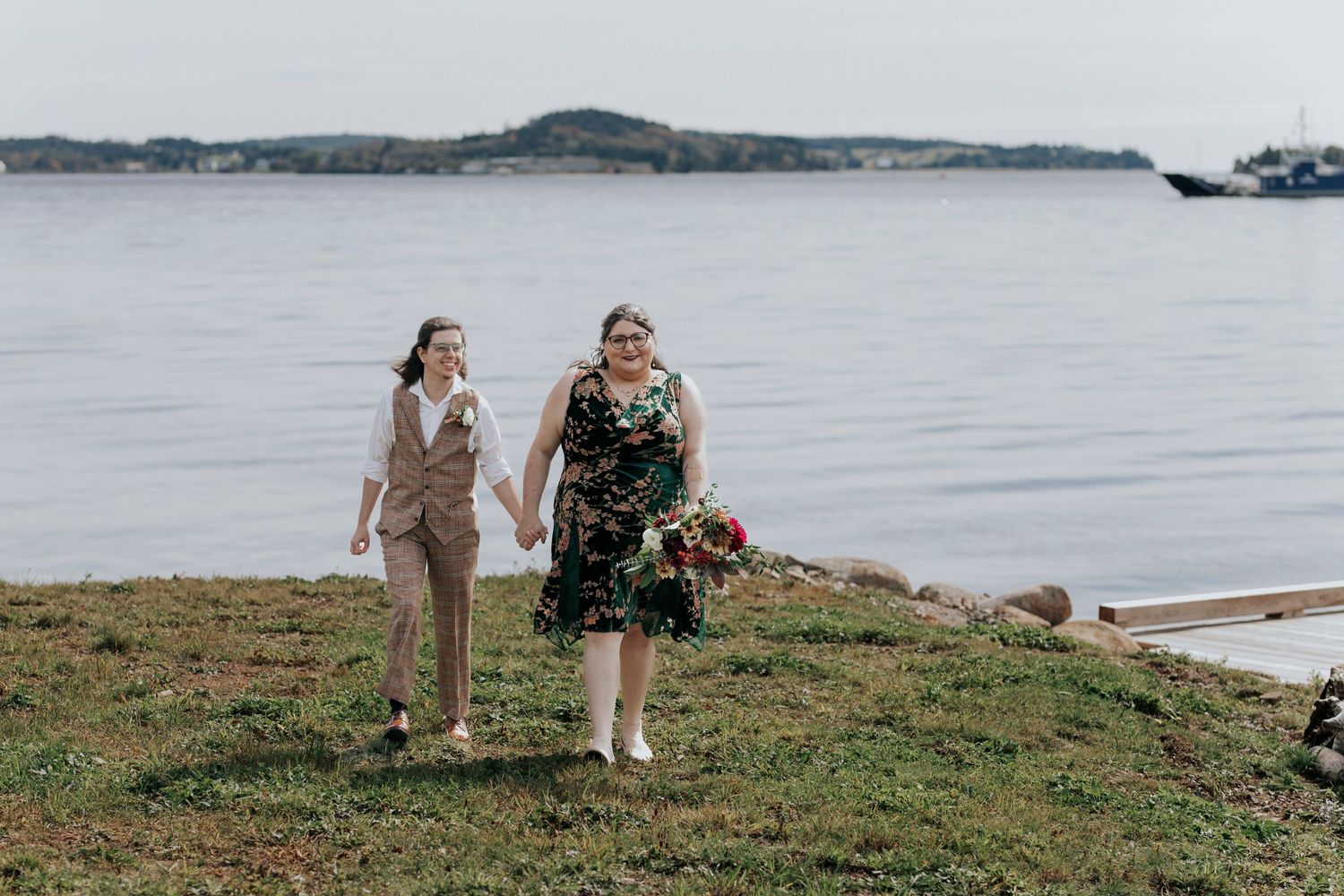 A wedding couple holding hands on a grassy hill overlooking a serene lake and distant mountains.