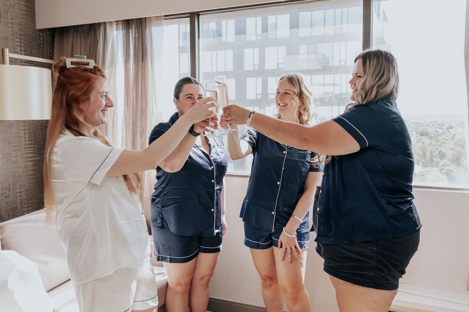 Four women in navy pajamas toast with champagne glasses in a bright hotel room during bridal prep.