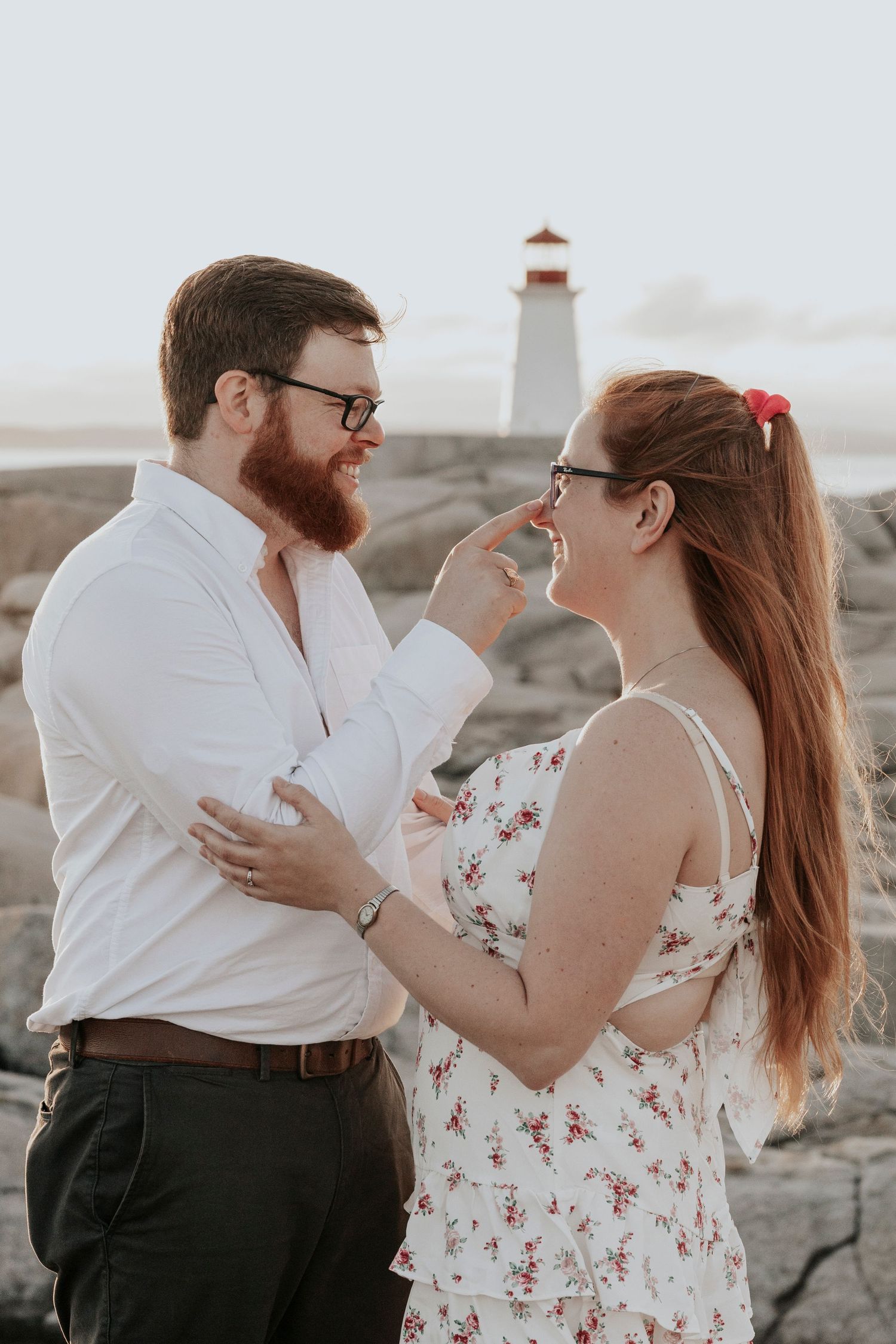 Bearded man gently touching smiling woman's face near Peggy's Cove lighthouse during golden hour engagement shoot.