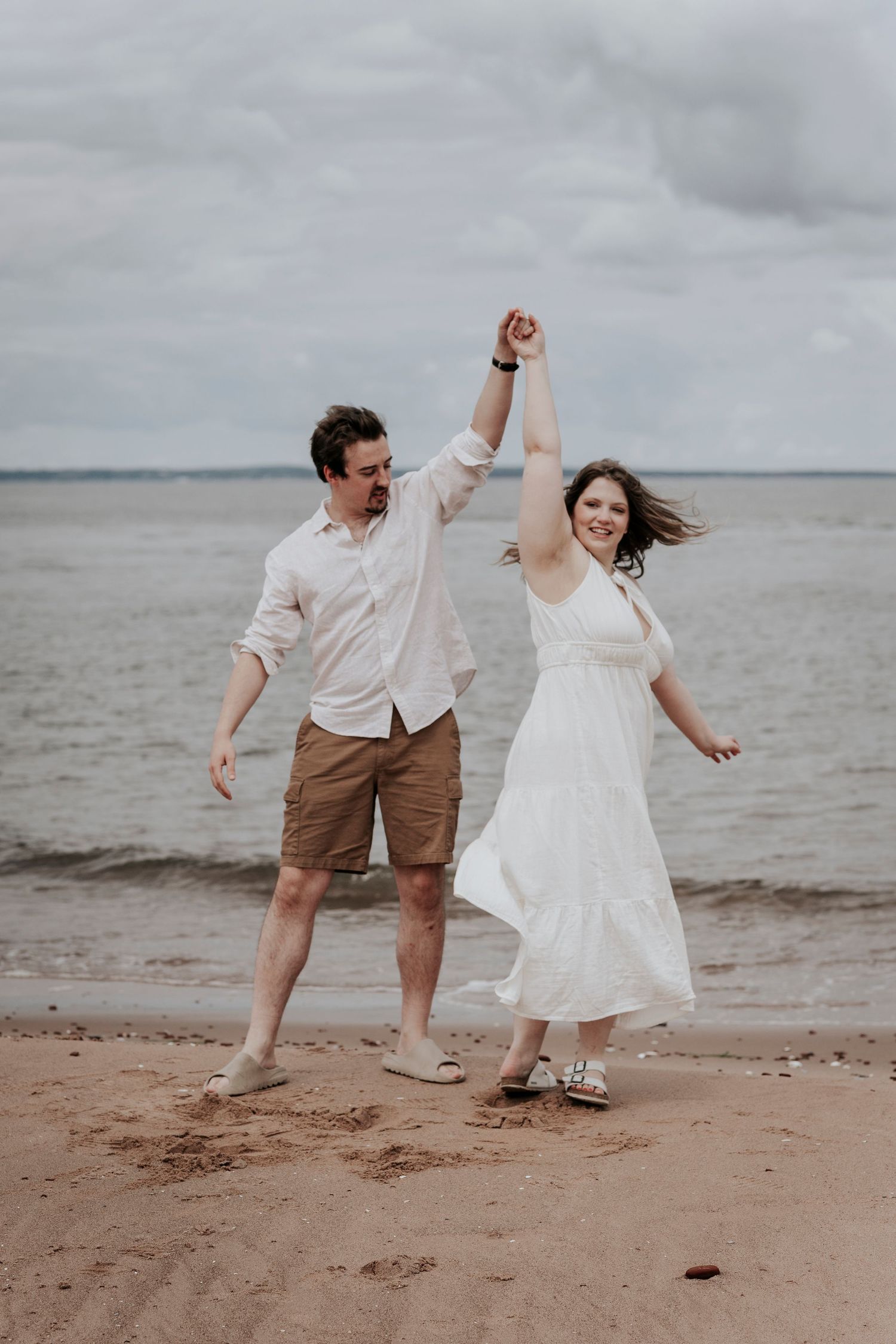 Joyful couple dancing and holding hands on sandy beach shore with cloudy sky and ocean backdrop.