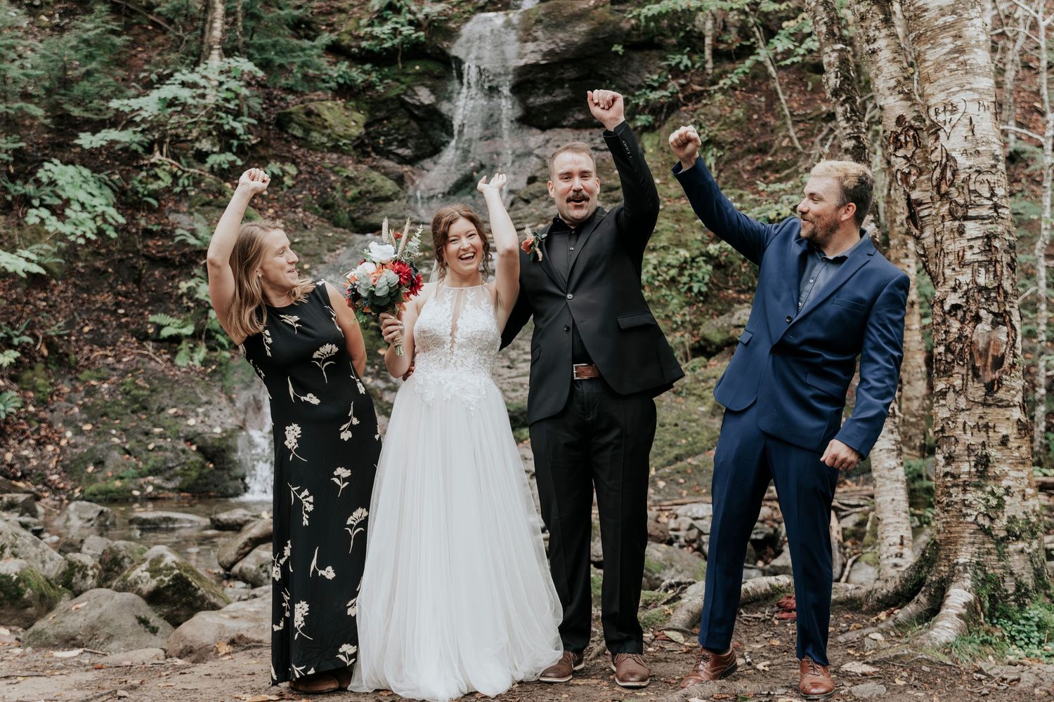 Joyful wedding party celebrating with raised fists in front of a waterfall in a lush forest setting.