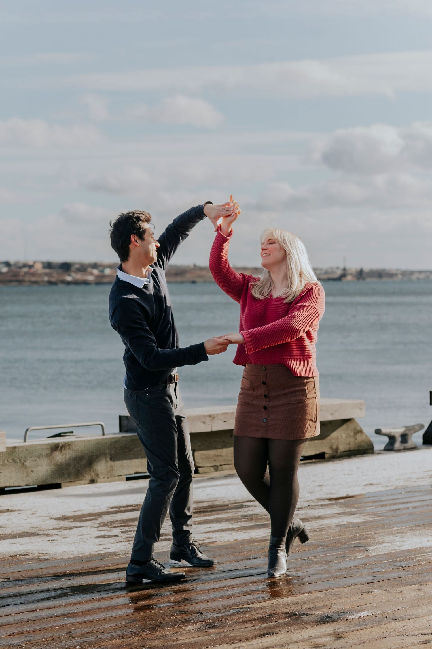 Couple dancing and spinning joyfully on a seaside pier under a cloudy sky.