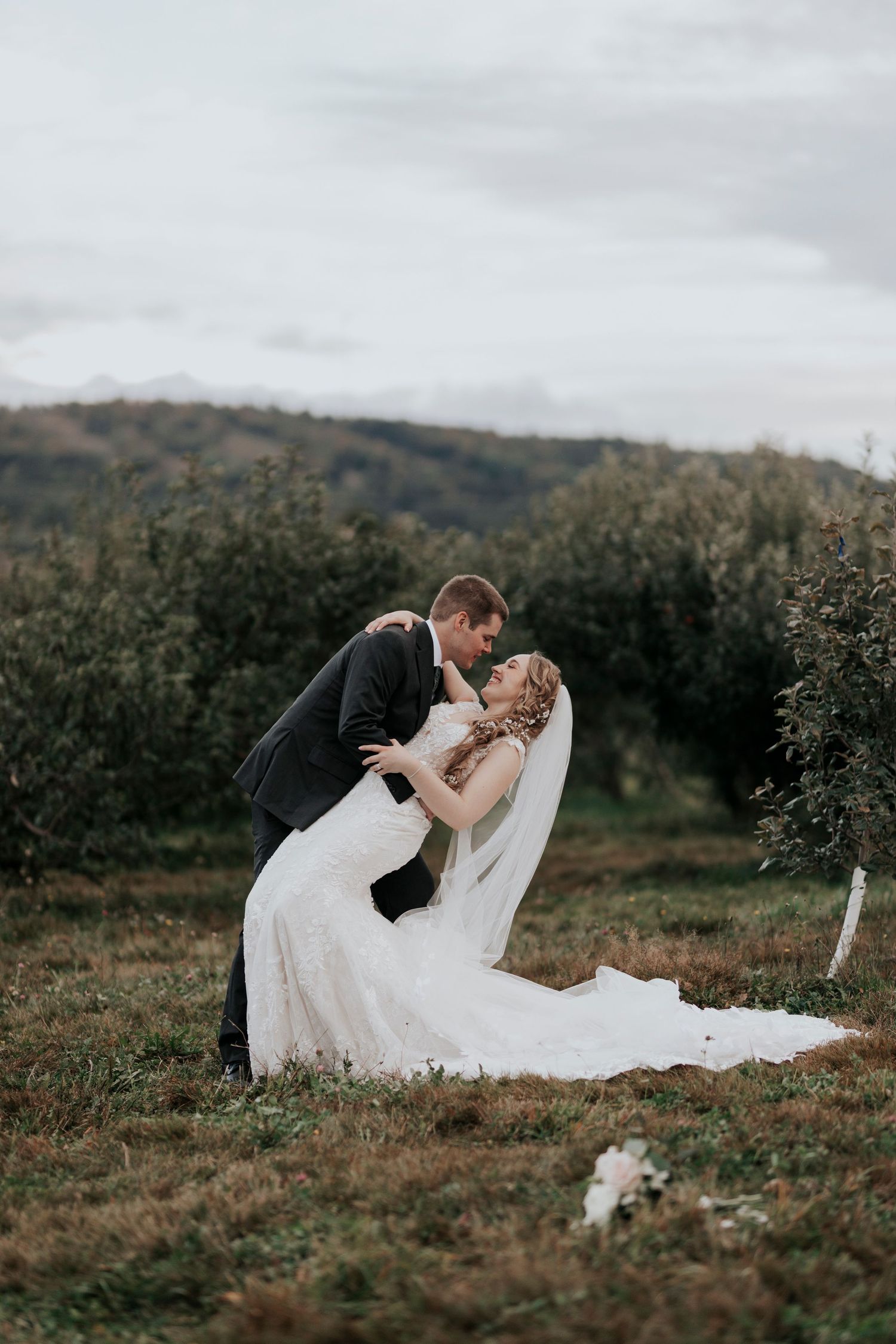 Bride and groom share a romantic dip kiss in a lush green orchard on their wedding day.