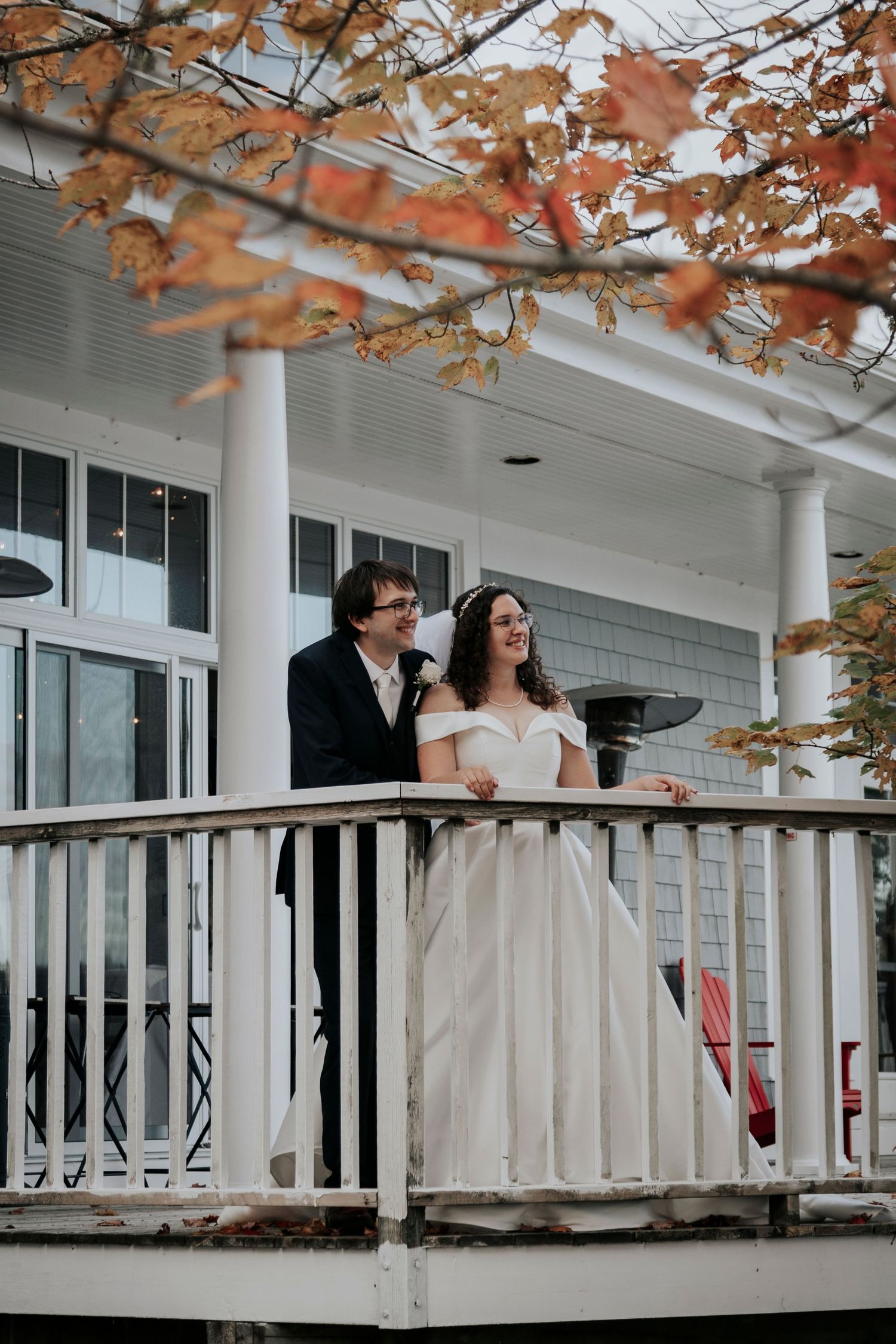 Bride and groom stand together on a white balcony framed by vibrant autumn orange leaves.