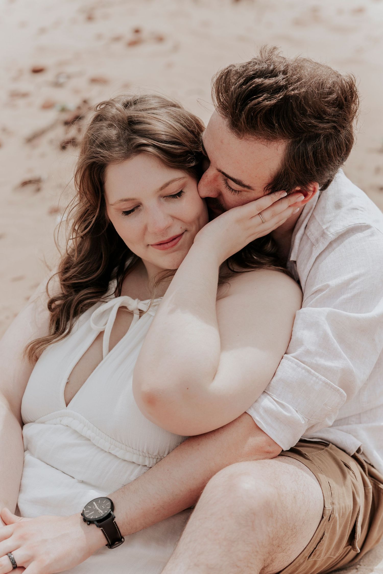 Couple sitting intimately on sandy beach as man tenderly kisses woman's cheek in close-up portrait.