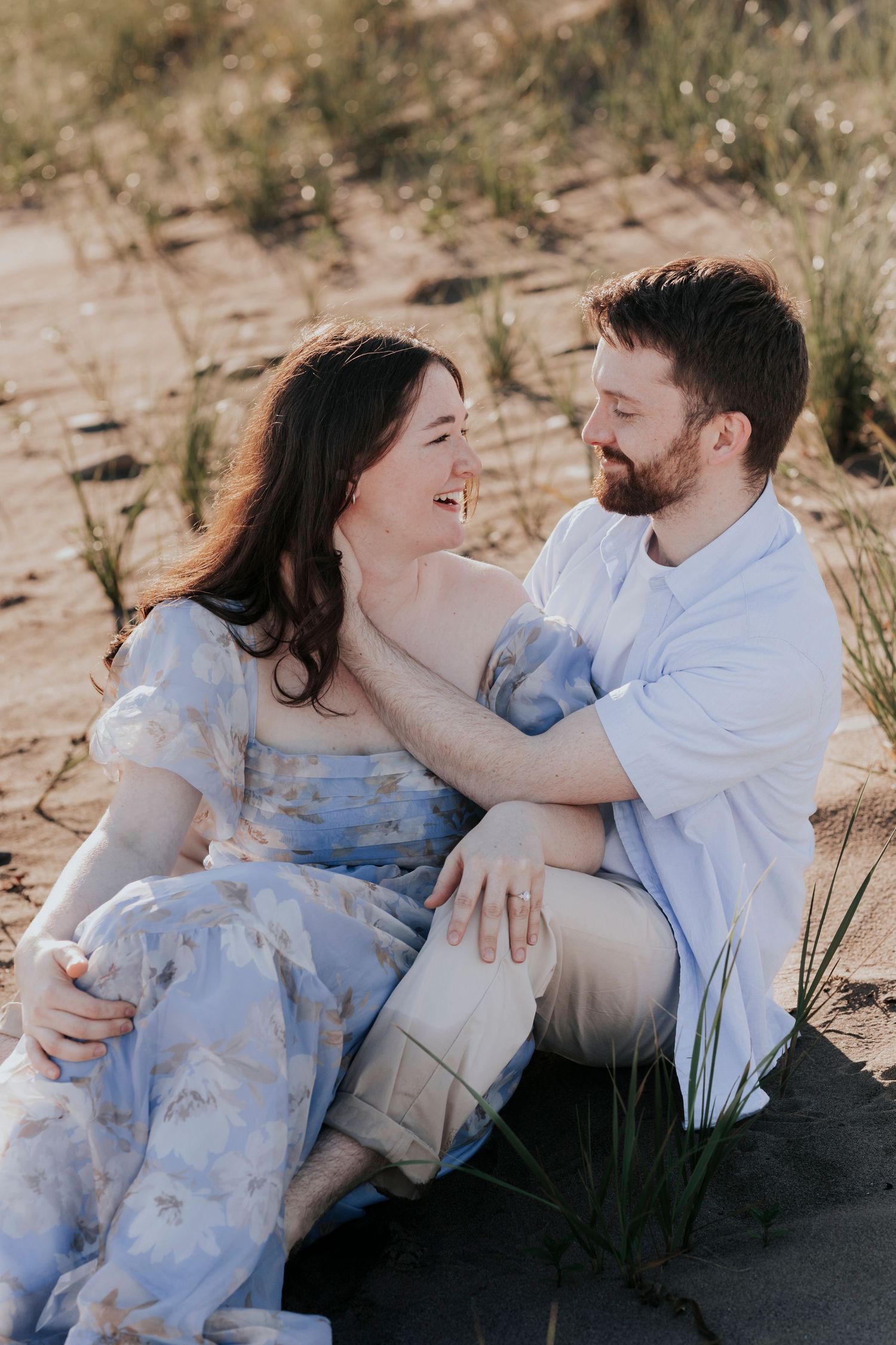 Smiling couple sitting together on sandy dunes surrounded by beach grass during golden hour.