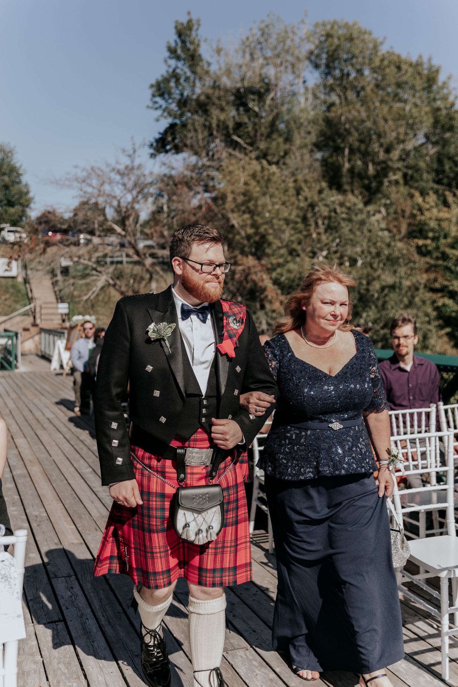 Groom in traditional Scottish kilt and black jacket walks down aisle escorted by woman in navy sequin dress.