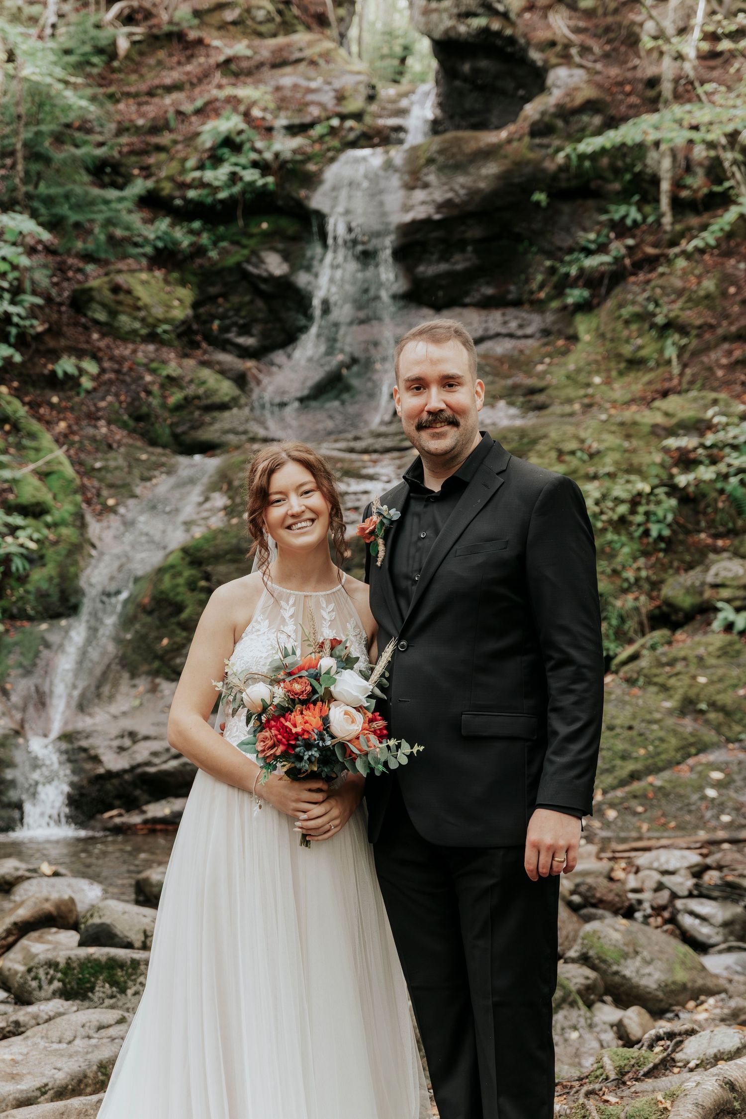 Smiling bride holding a vibrant red and white bouquet posing with groom before a woodland waterfall.