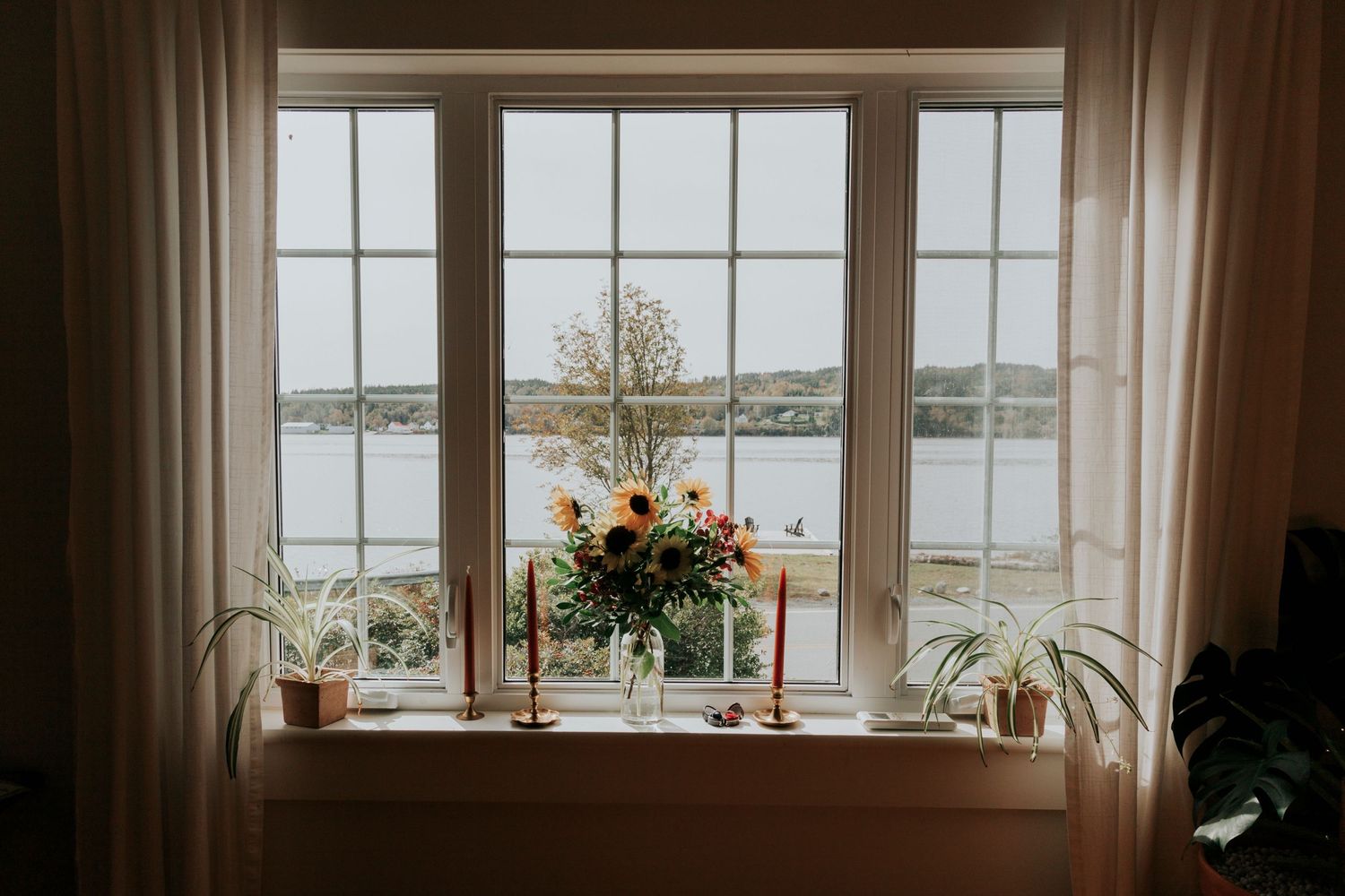 A floral arrangement sits on a windowsill overlooking a serene lake view through a large paned window with beige curtains.