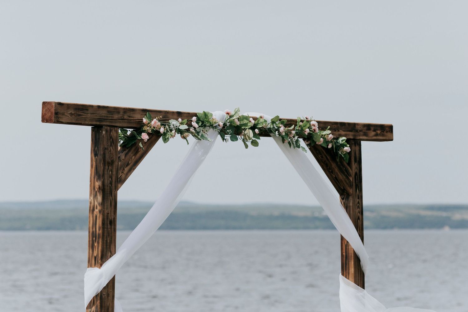 Rustic wooden wedding arch decorated with white fabric and greenery overlooking Bras D'Or in Cape Breton, NS
