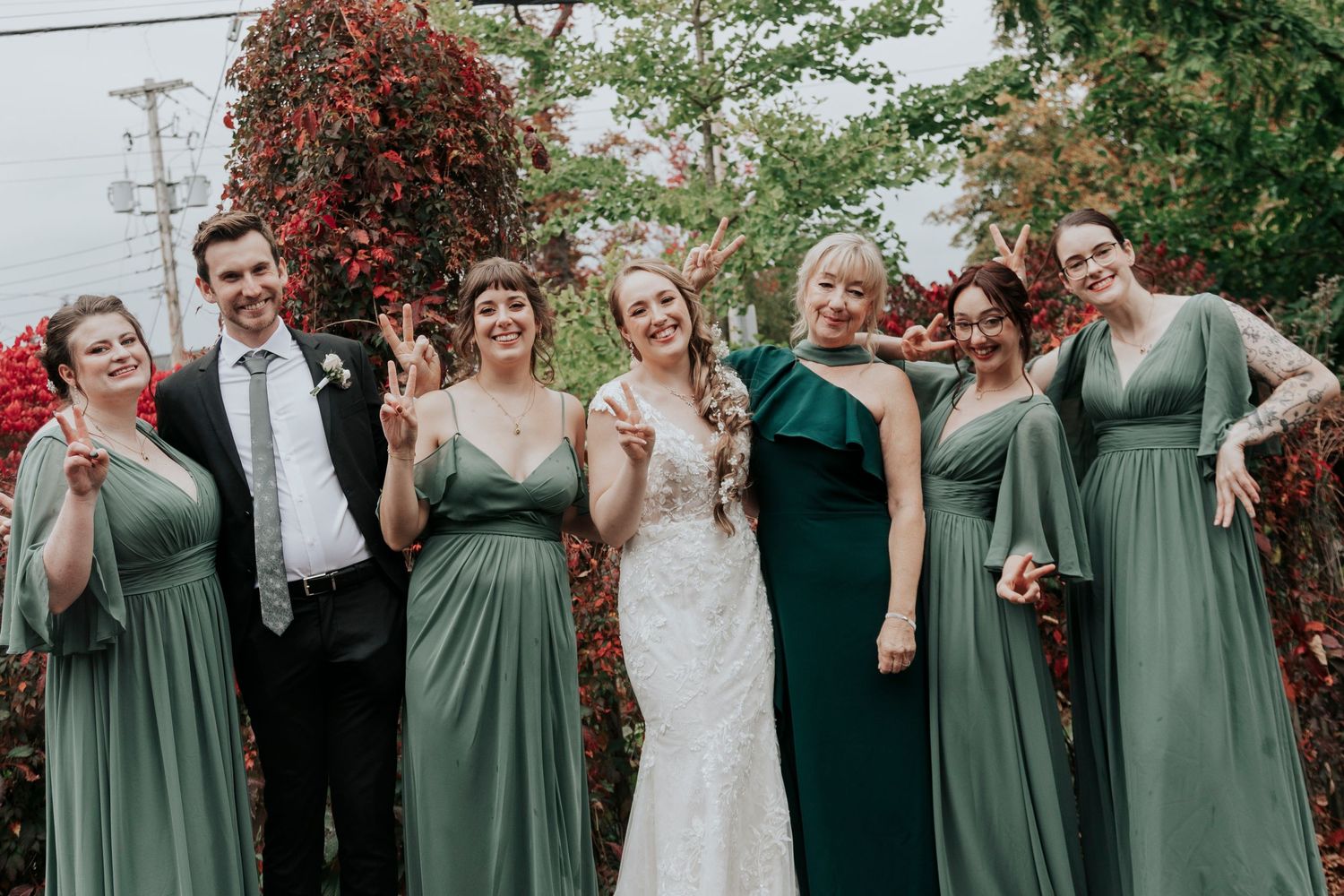 Wedding party in sage green dresses poses outdoors with bright autumn foliage.