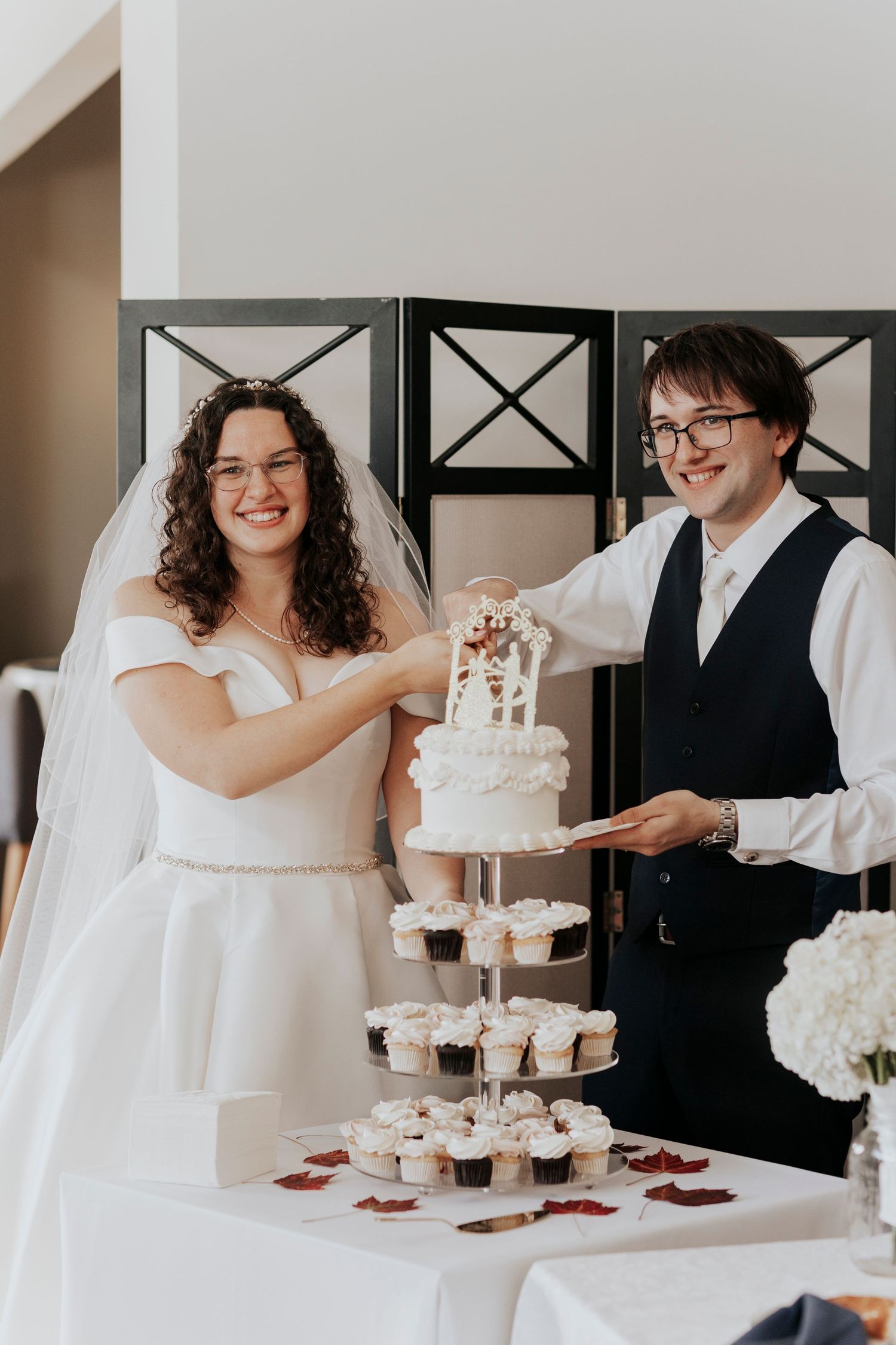 A happy couple sharing a special moment while cutting their white tiered wedding cake with cupcakes at their reception.