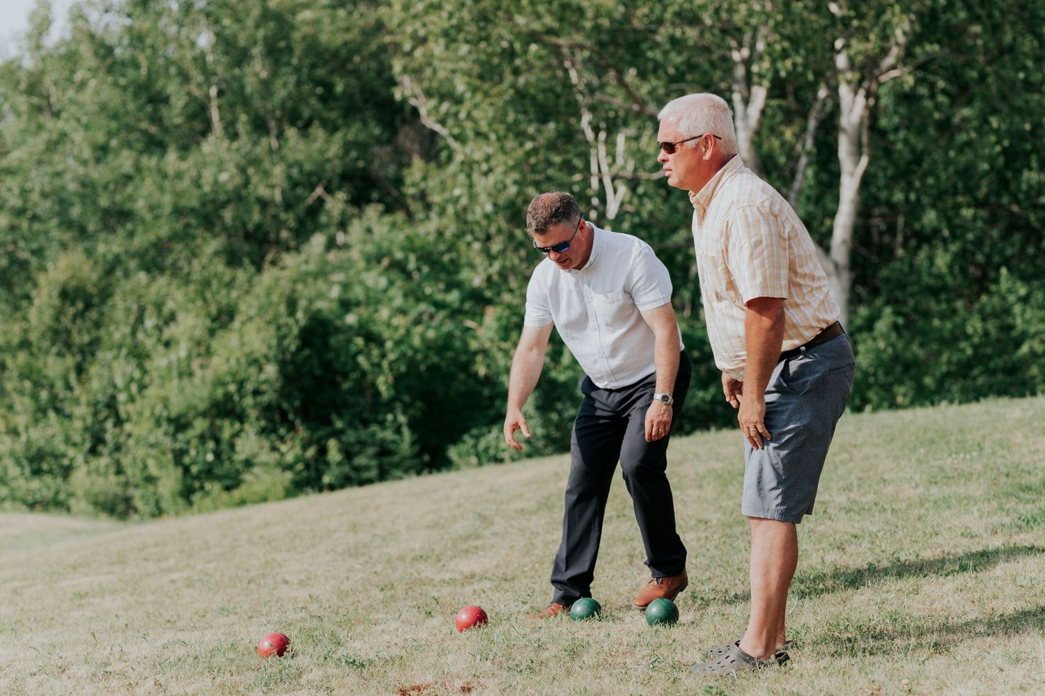 Two people playing bocce ball on a grassy hillside with trees in the background on a sunny day.