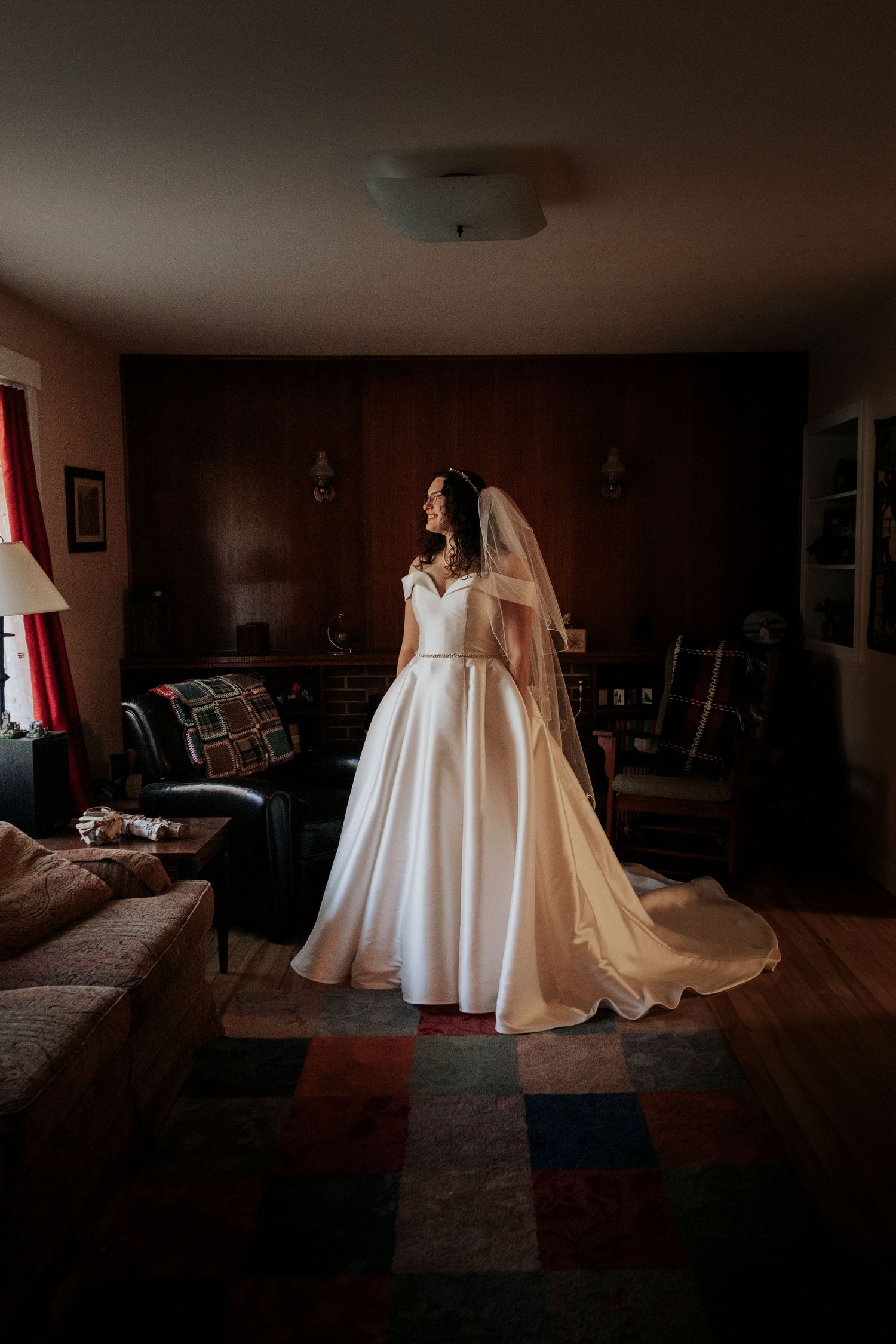 A bride stands in a dimly lit living room wearing an elegant white satin ball gown with a flowing veil and train.