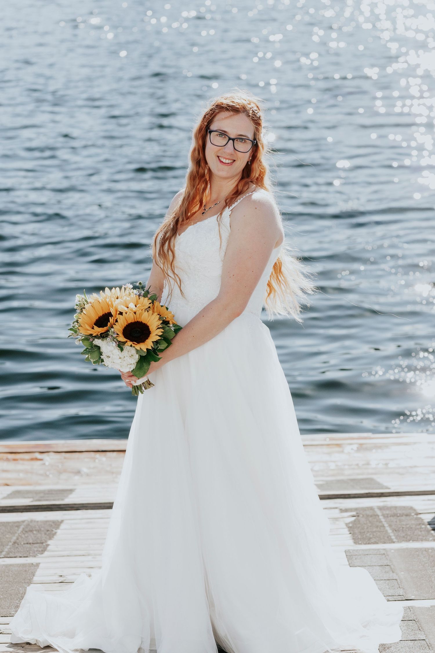 Smiling bride in white gown holding sunflower bouquet standing on dock by sparkling water.