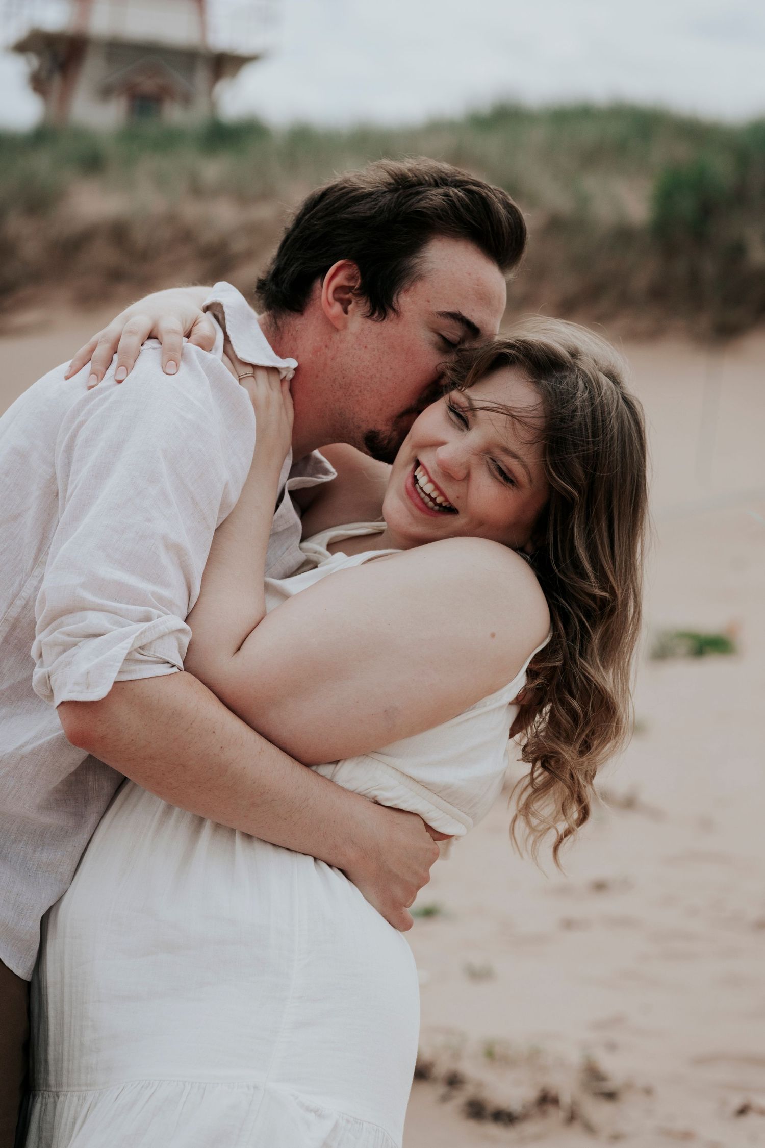 Happy couple embracing on beach, man kissing woman's cheek as she laughs joyfully in white dress.