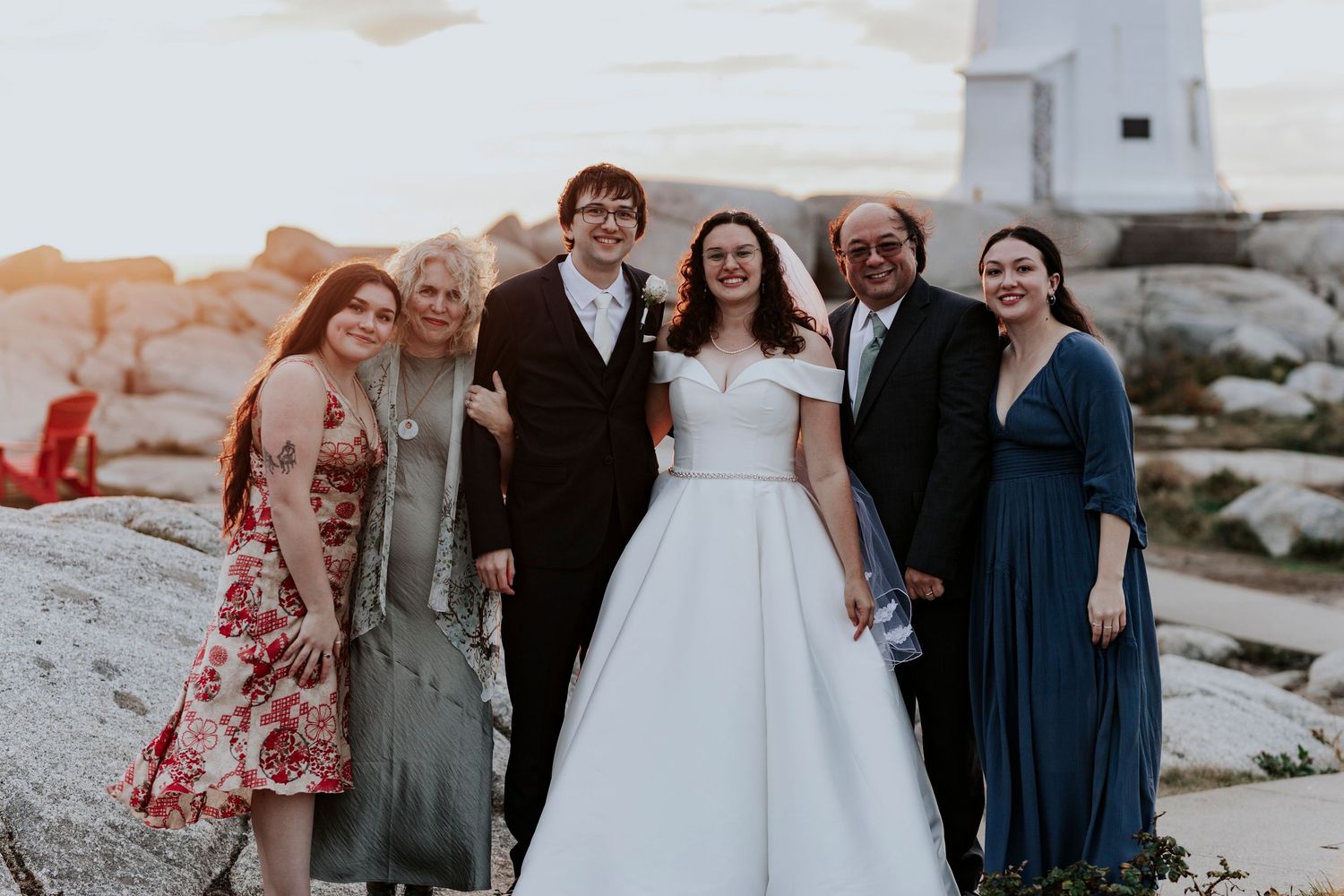 A group of people in formal attire gather on coastal rocks at sunset with the woman at center in a white wedding dress.