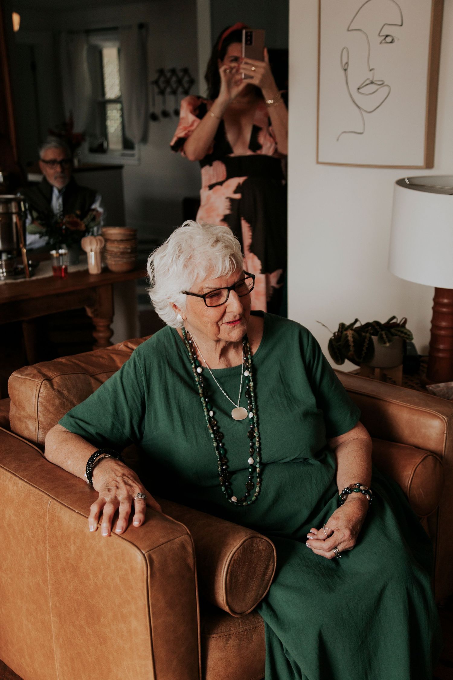 An elderly woman in a green dress and glasses sits on a brown leather couch with a minimalist line drawing on the wall behind.