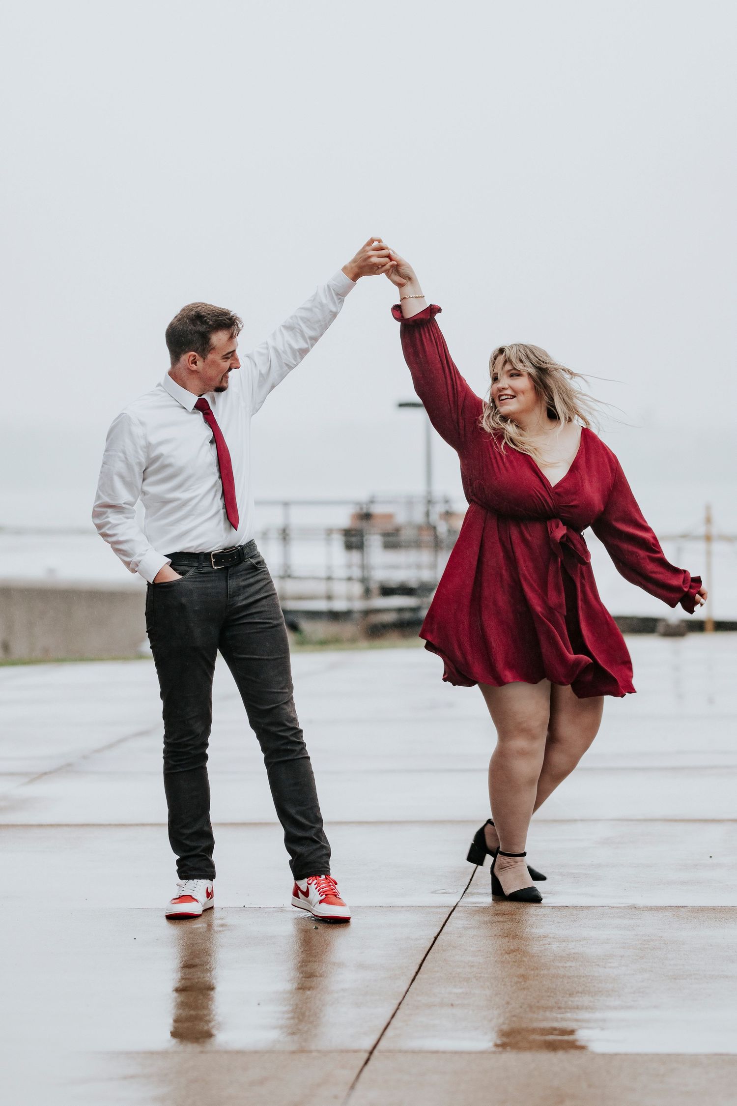 Joyful couple dances in the rain on a wet pier, man spinning woman in red dress.