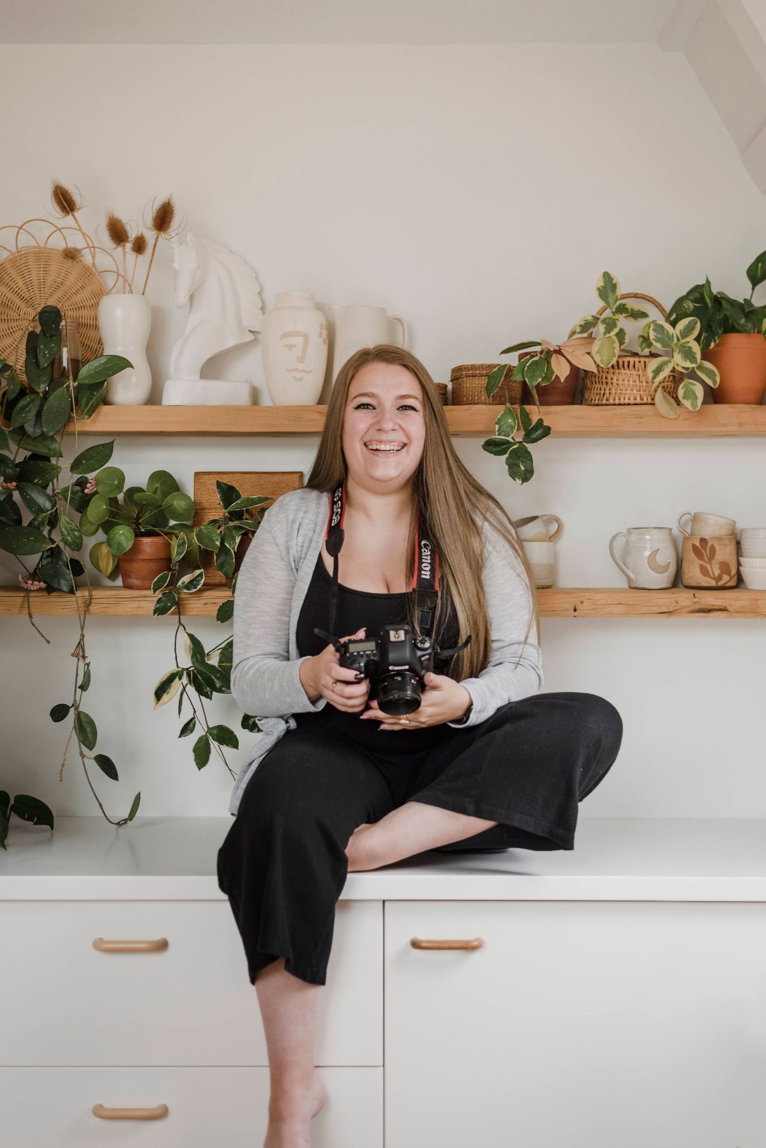Smiling female photographer sitting cross-legged holding camera, surrounded by plants and shelves.