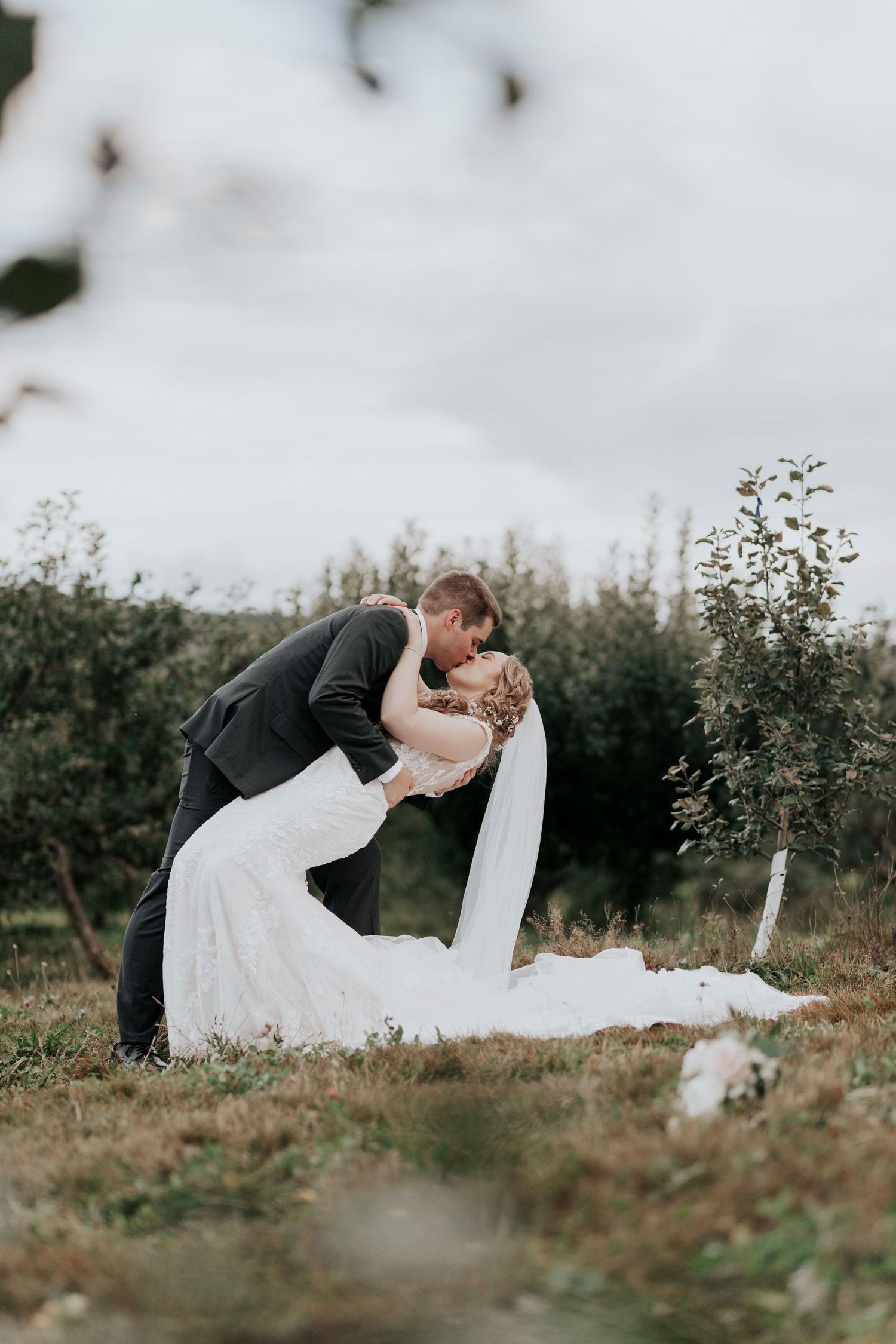 Bride and groom share a romantic dip kiss in a lush green orchard on their wedding day.
