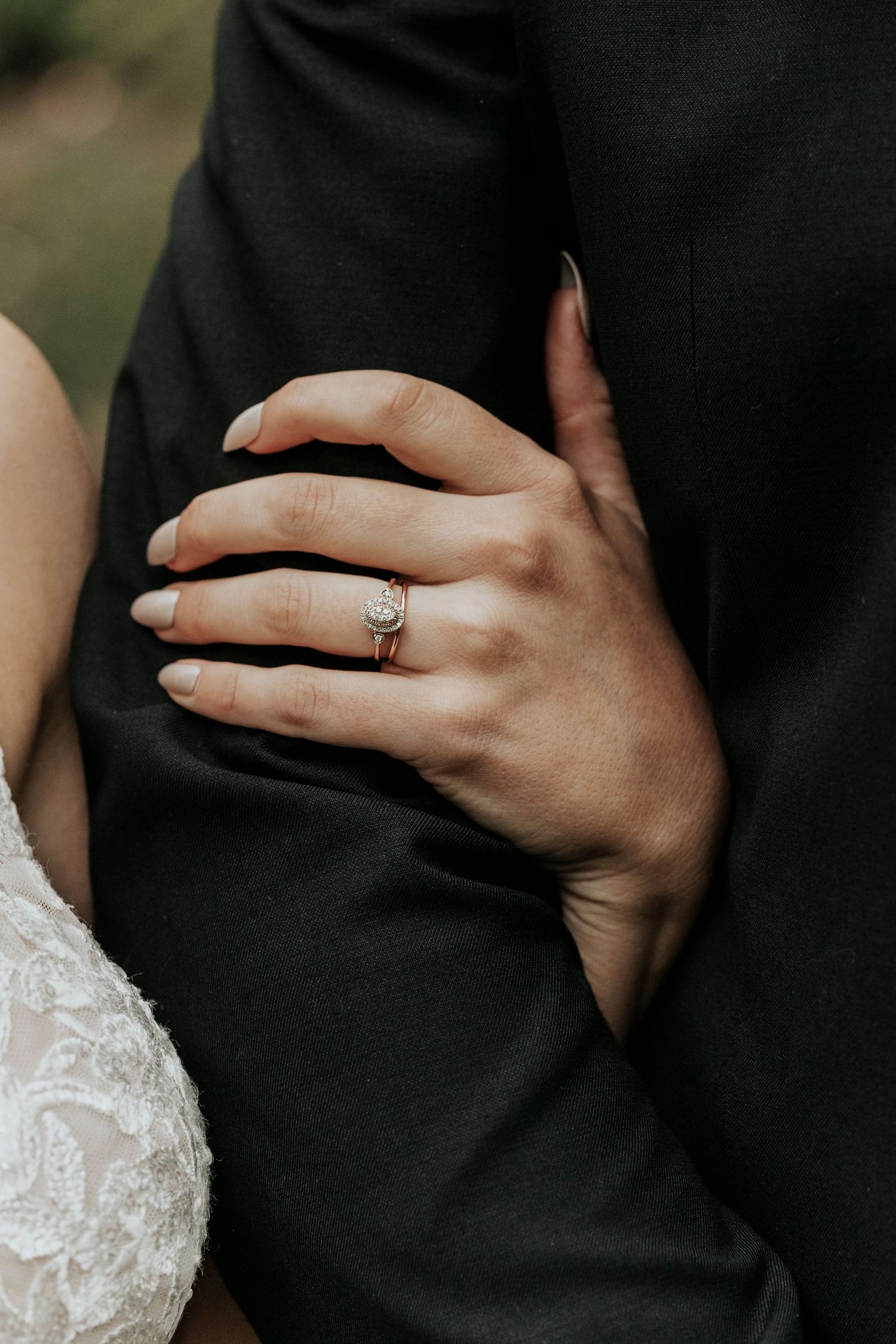 Close-up of bride's hand with elegant diamond engagement ring resting on groom's black suit jacket.