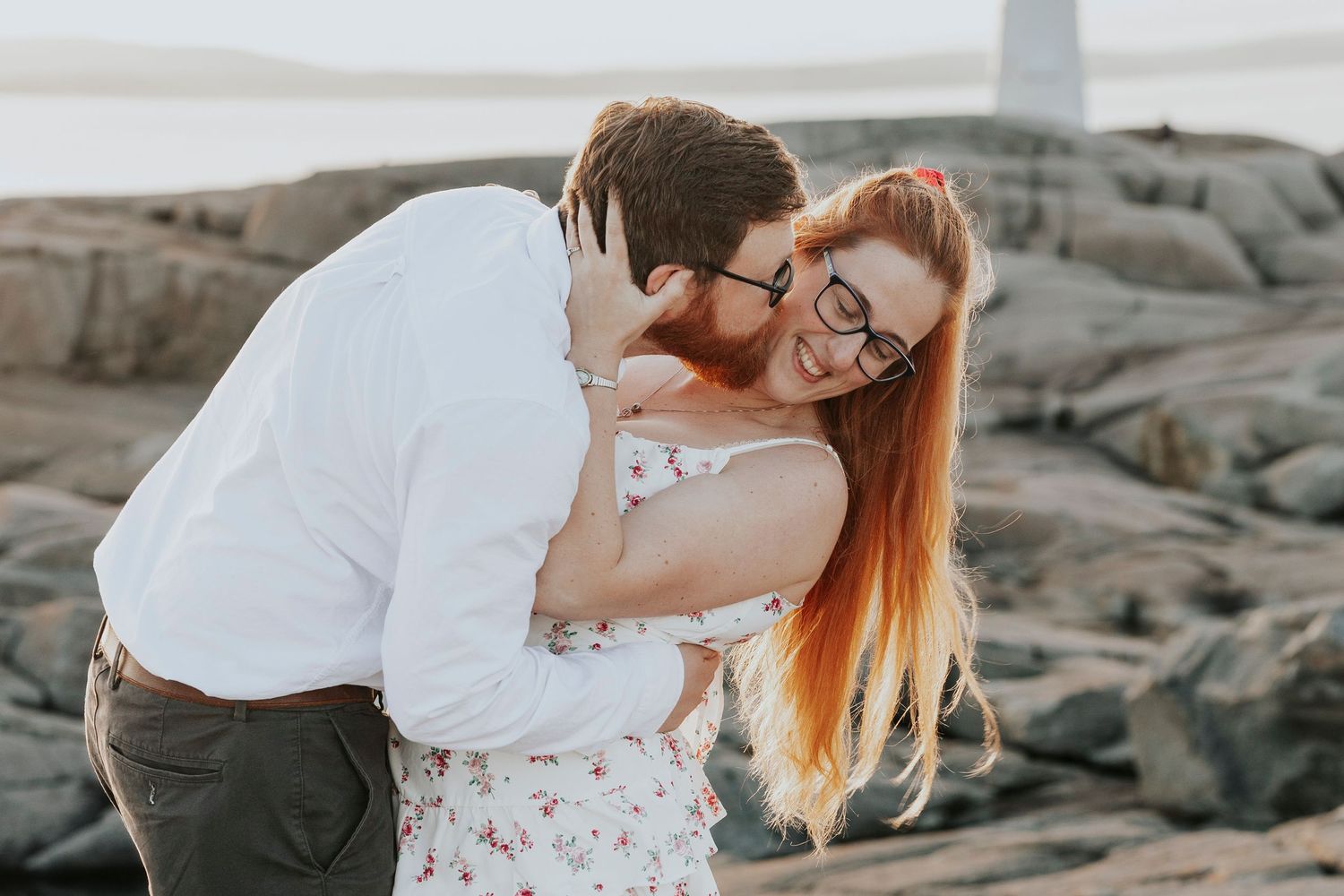Woman laughing joyfully as man nuzzles her neck during romantic couples photo session on rocky seashore.