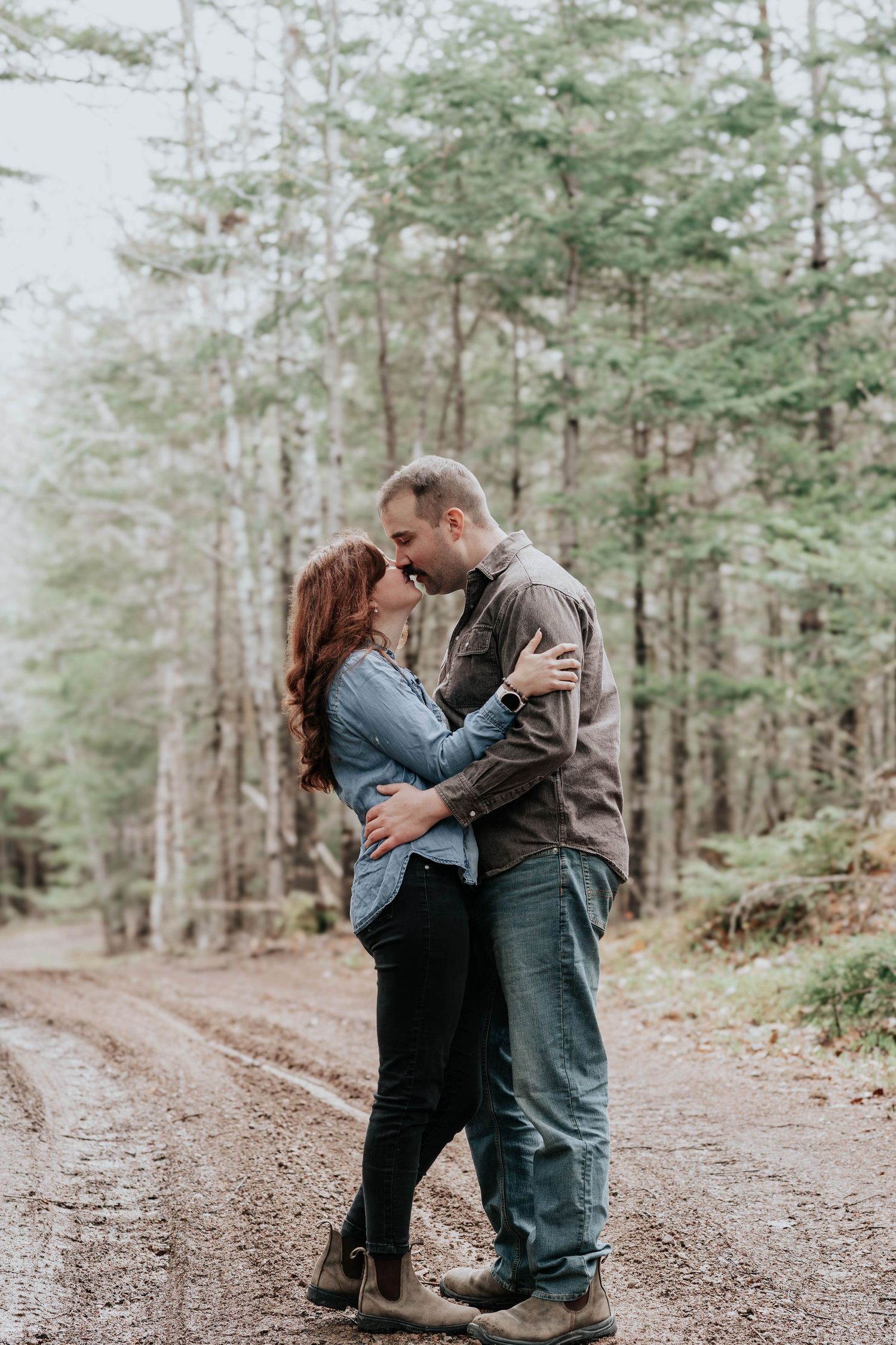 Couple sharing a kiss on a misty forest dirt road surrounded by tall evergreen trees.
