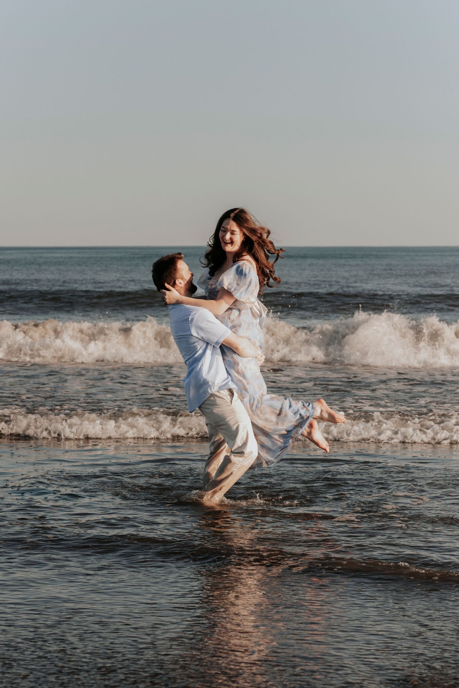 Man lifting woman in his arms while laughing in ocean waves during a romantic beach photoshoot.