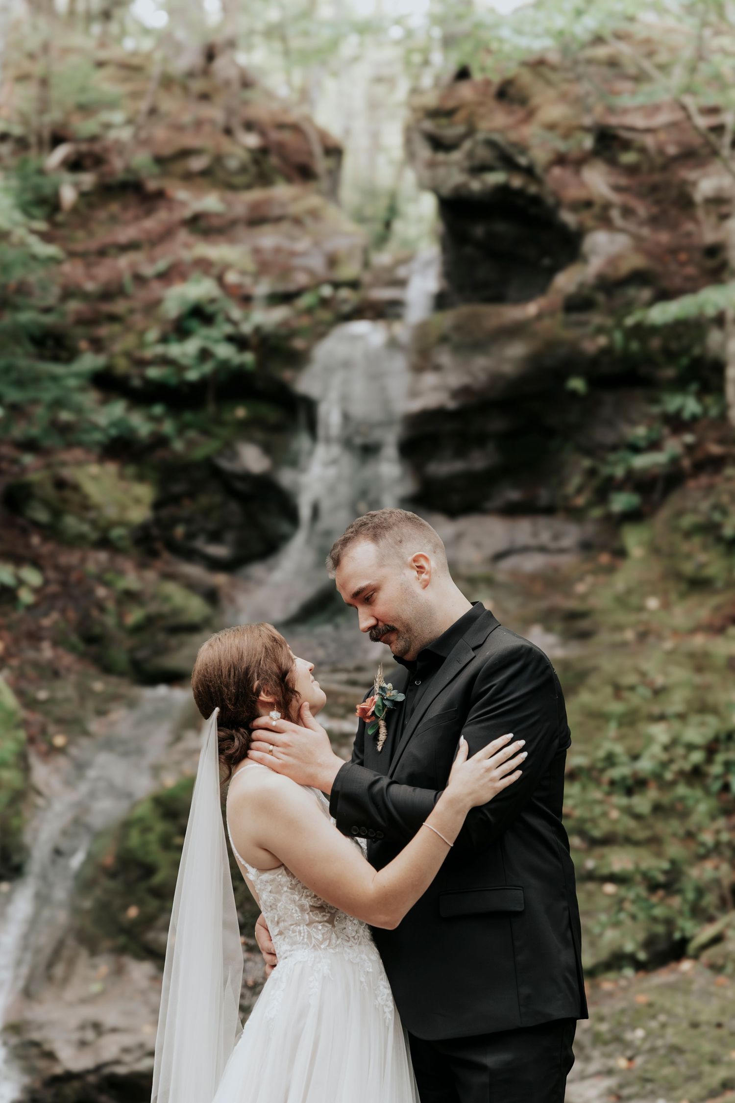 Bride and groom embracing tenderly in front of a lush waterfall in a forest setting.