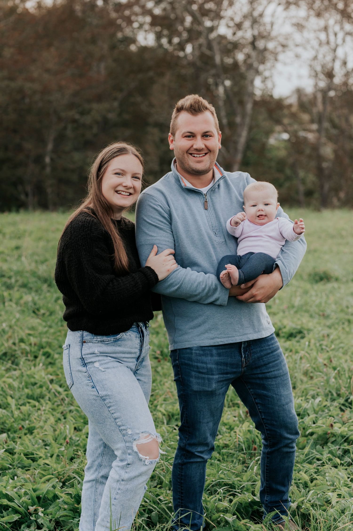 Happy young family with mother, father, and baby posing in a green field during a fall outdoor photo session.