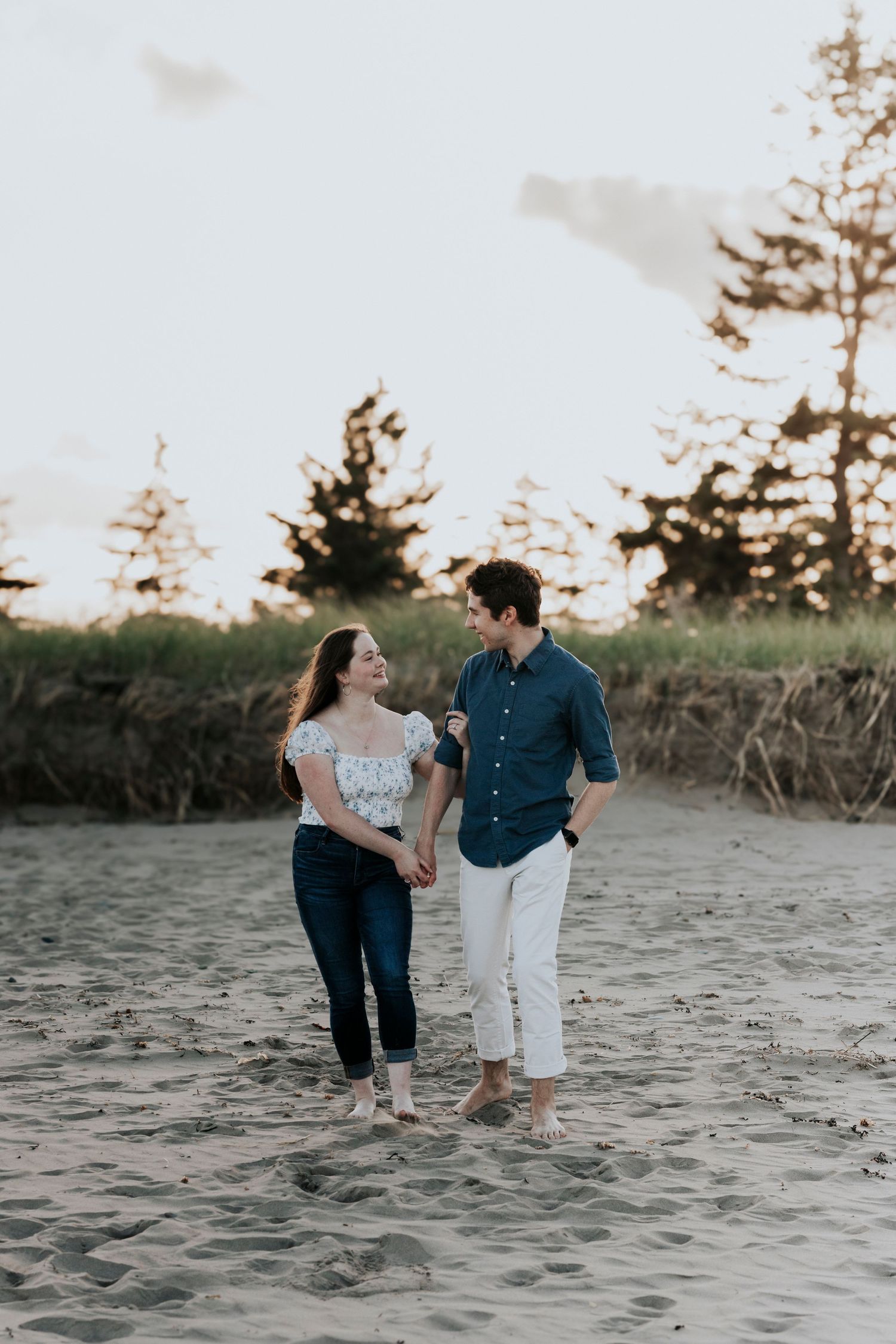 Romantic couple walking hand in hand on sandy beach at golden hour with trees in background.