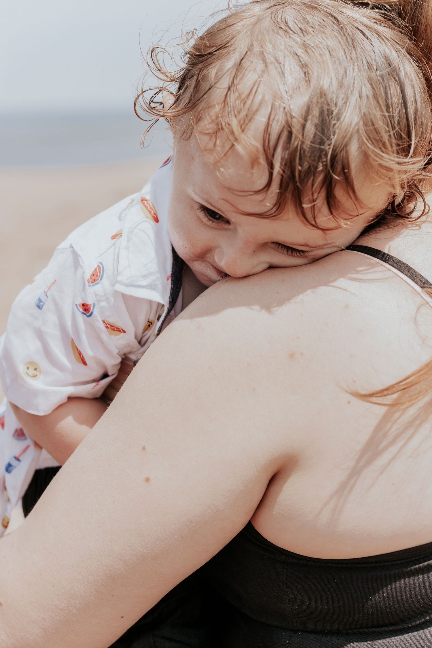 Wet toddler resting head on mothers shoulder at the beach after playing in water.