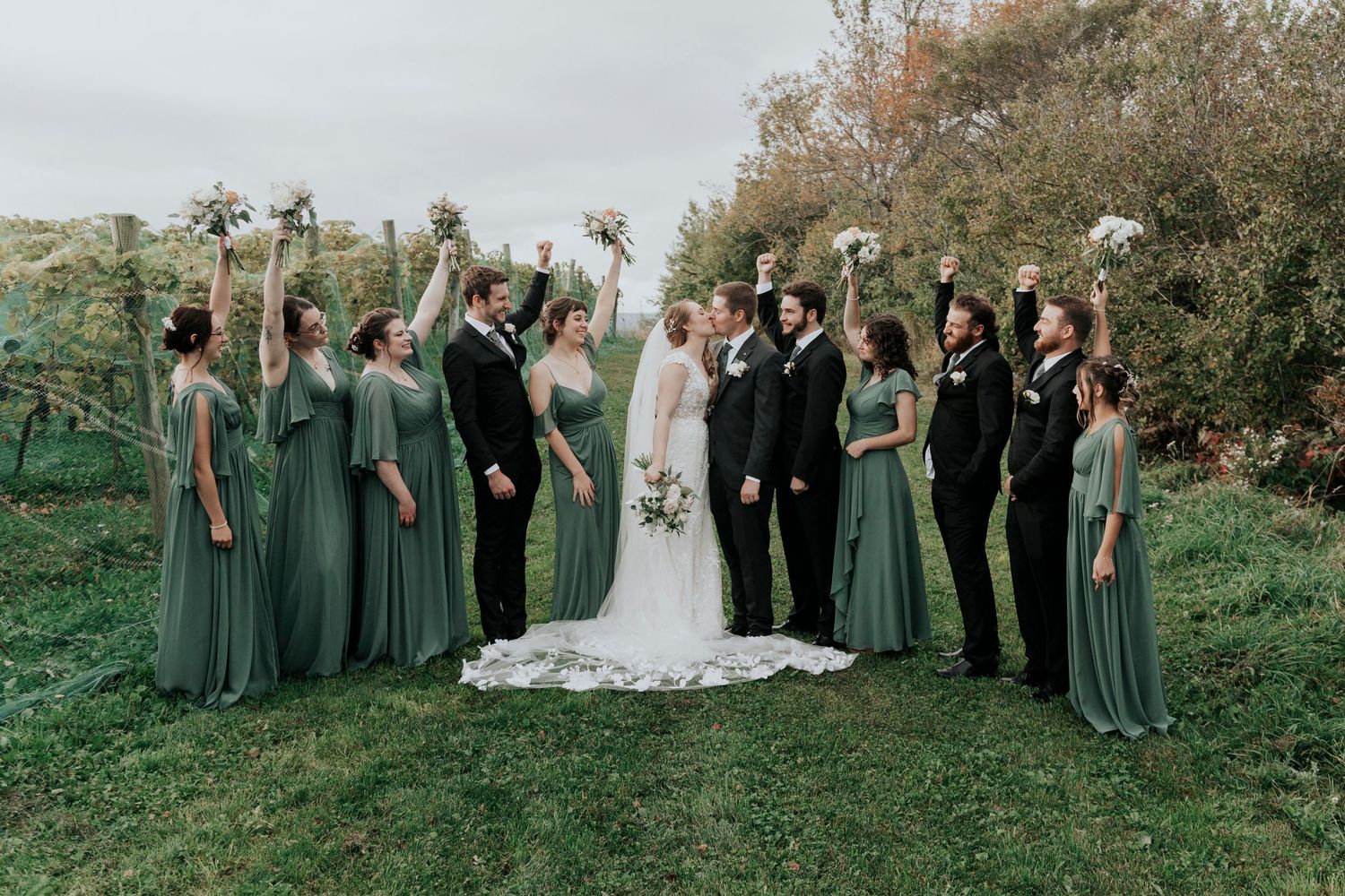 Wedding party in sage green dresses and black suits posing on grassy hillside.