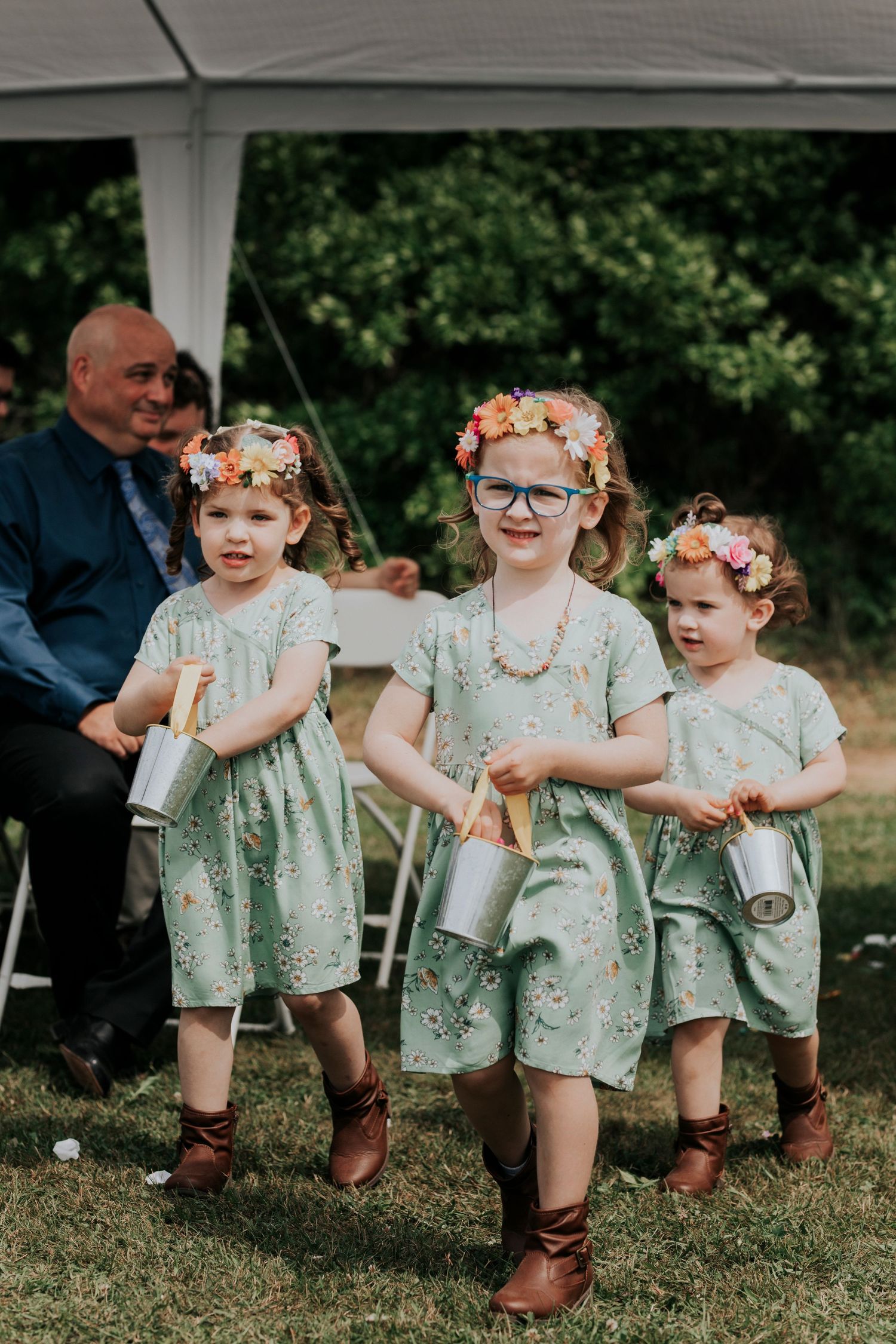 Three young flower girls in matching mint green dresses and brown boots walk outdoors under a white tent at a formal event.