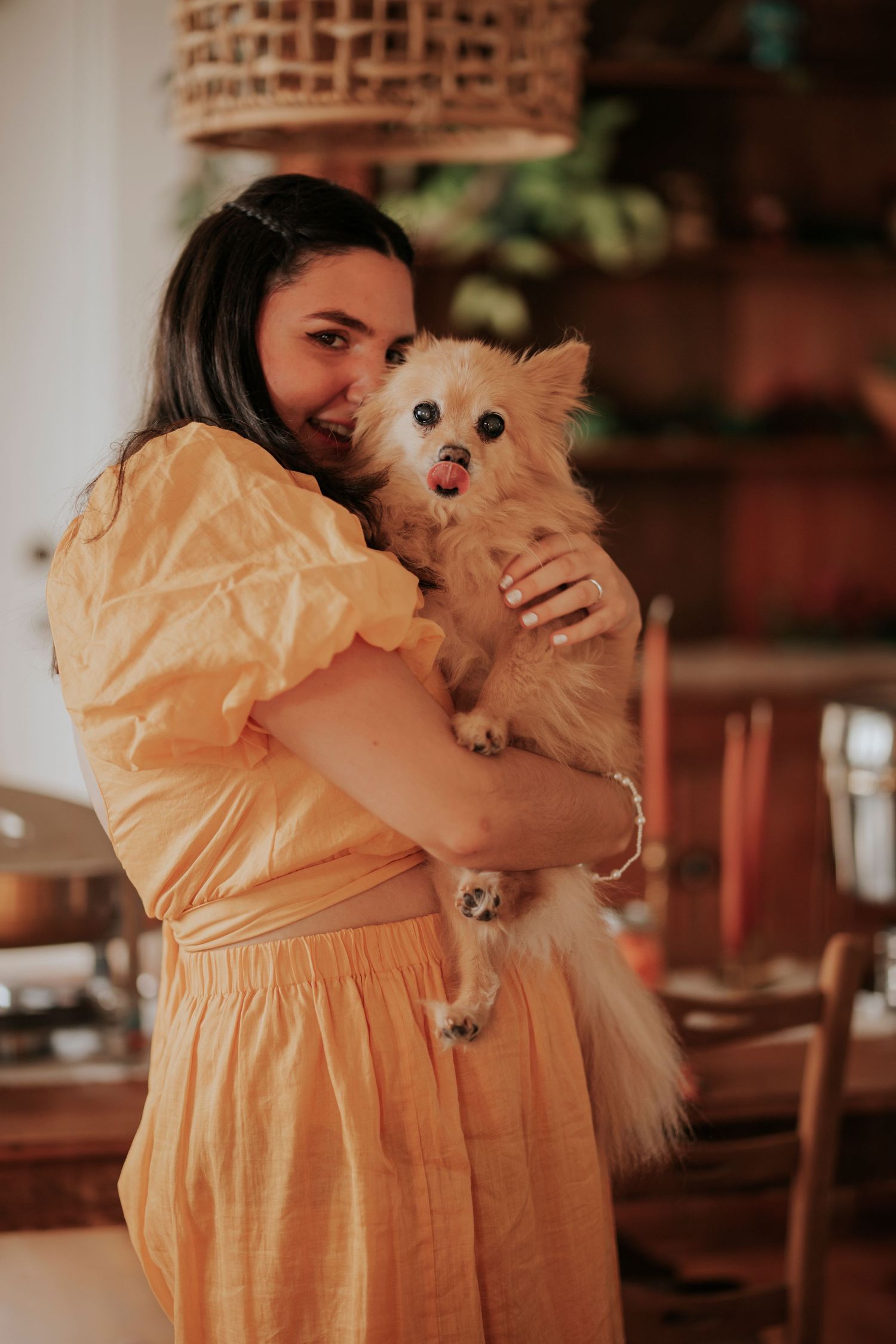 A long-haired Pomeranian puppy is held lovingly by someone wearing a yellow summer dress in a warm indoor setting.