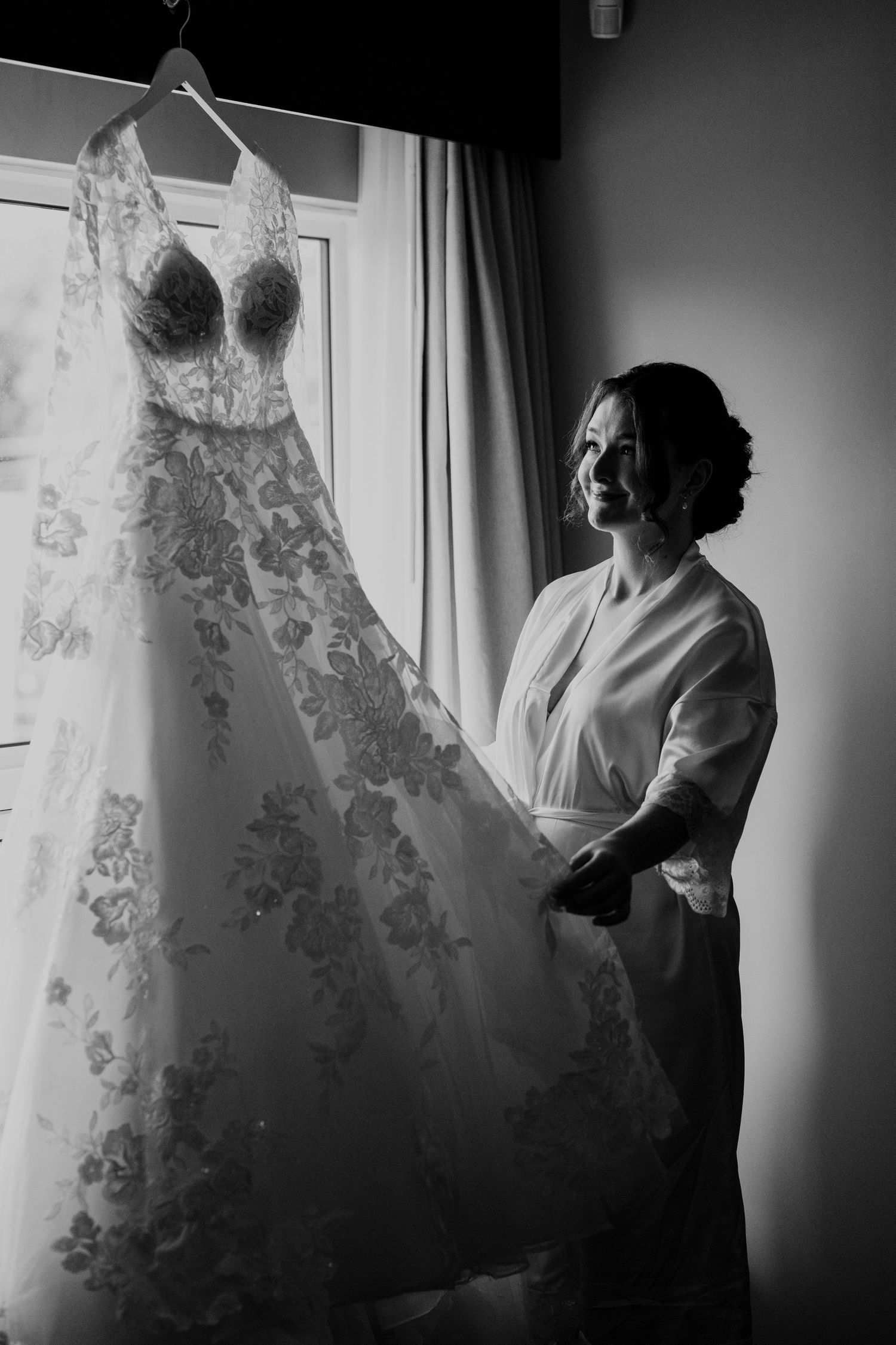Black and white photo of bride admiring her lace wedding gown hanging by a window.