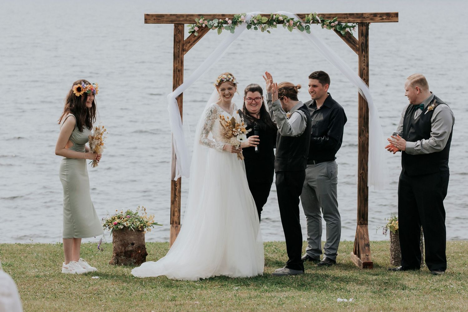 An outdoor wedding ceremony taking place by a lake under a rustic wooden arch with attendees gathered together.