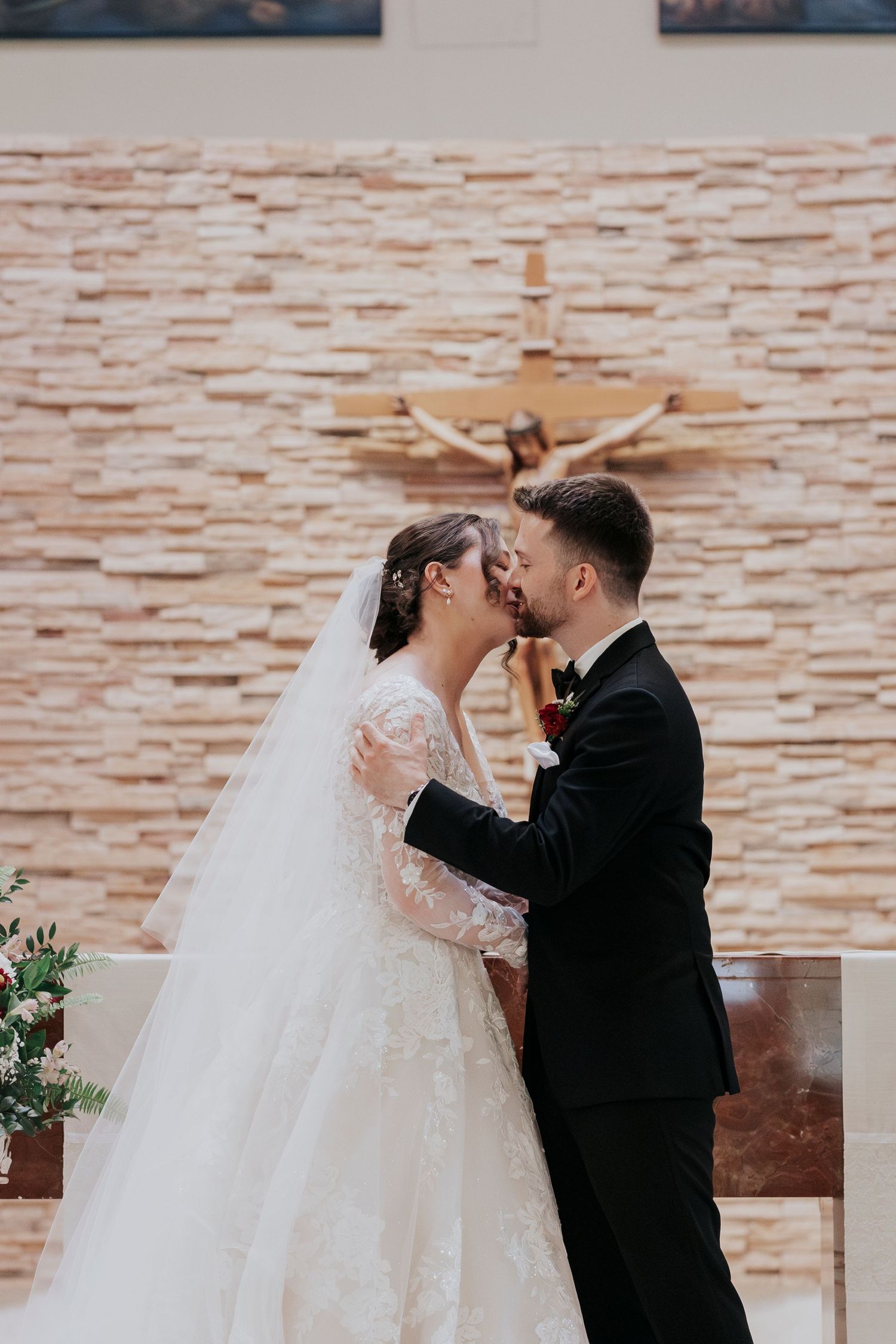 A romantic wedding moment in front of a textured stone wall with a cross.