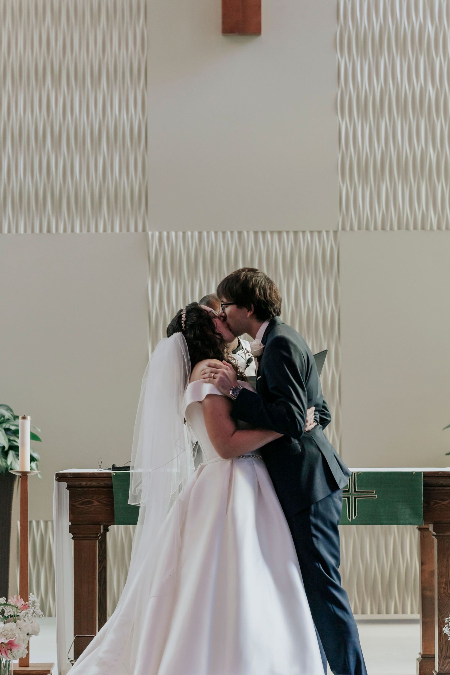 Bride and groom sharing first kiss at church altar in front of cross on wedding day.