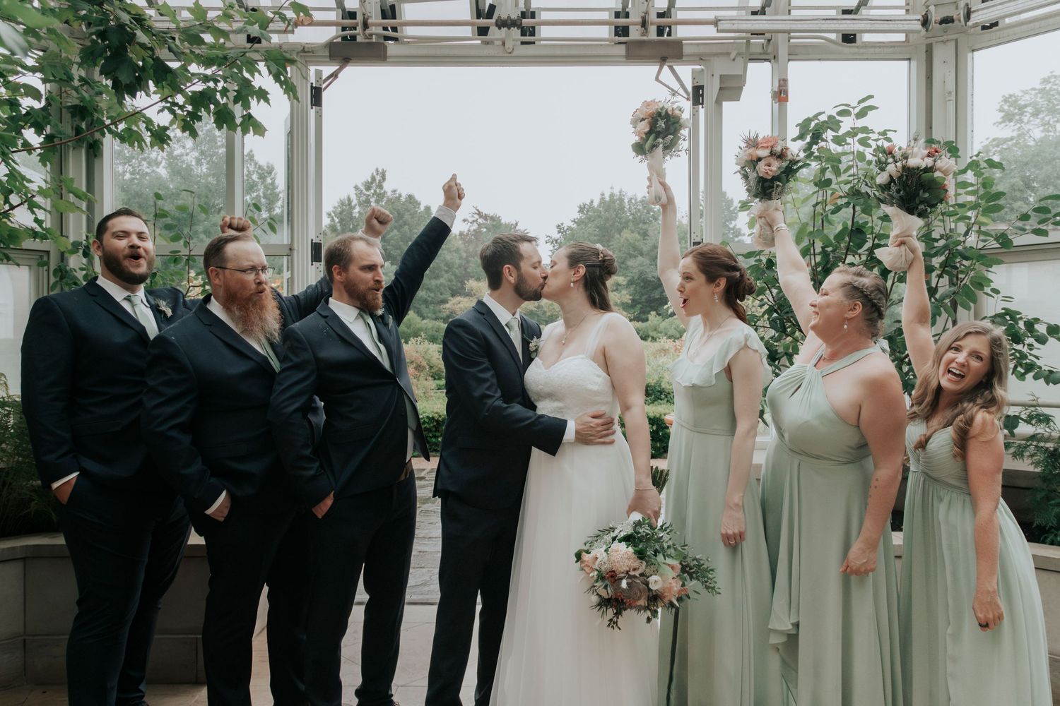 Wedding party celebrating in a greenhouse venue with bridesmaids in sage green dresses and groomsmen in navy suits.